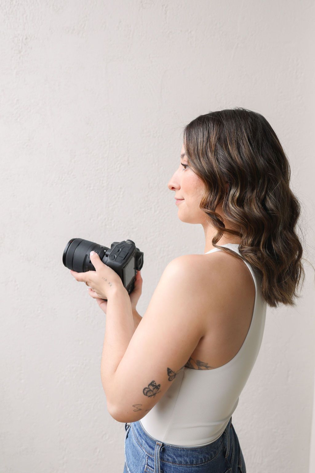 A person with long, wavy hair holds a camera up to their eye, ready to take a photo. They are wearing a sleeveless top a