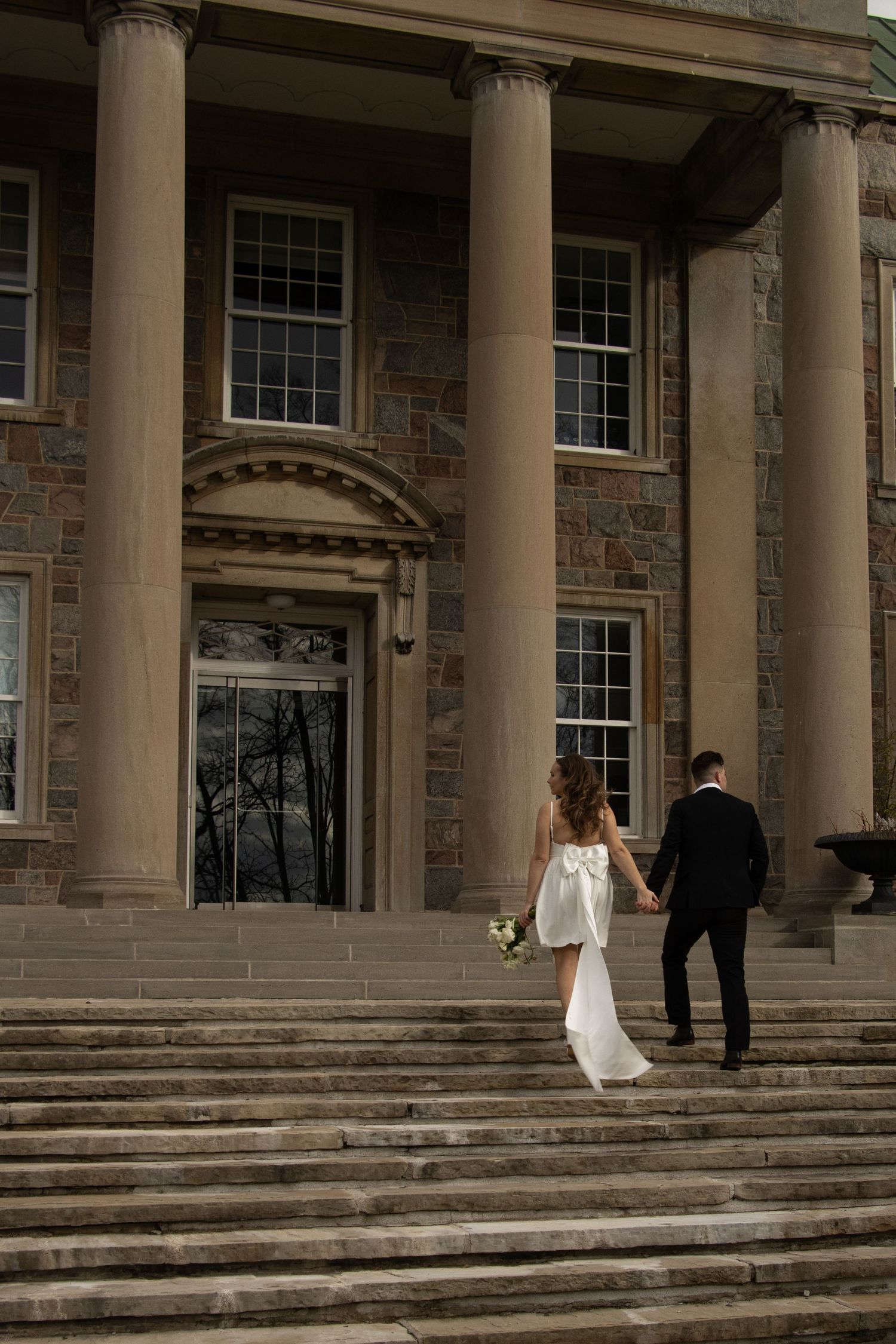 A bride in a white dress and a groom in a black suit ascend stone steps towards a building with large pillars and glass 