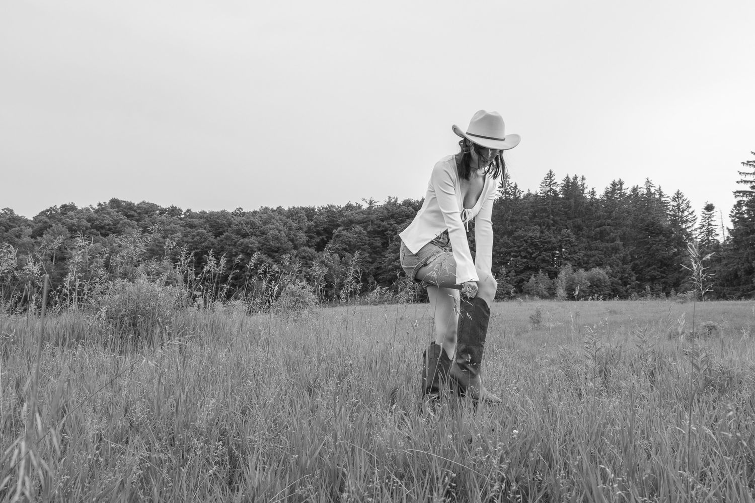 A black-and-white photo of a person in a cowboy hat and boots standing in a grassy field. The person appears to be adjus