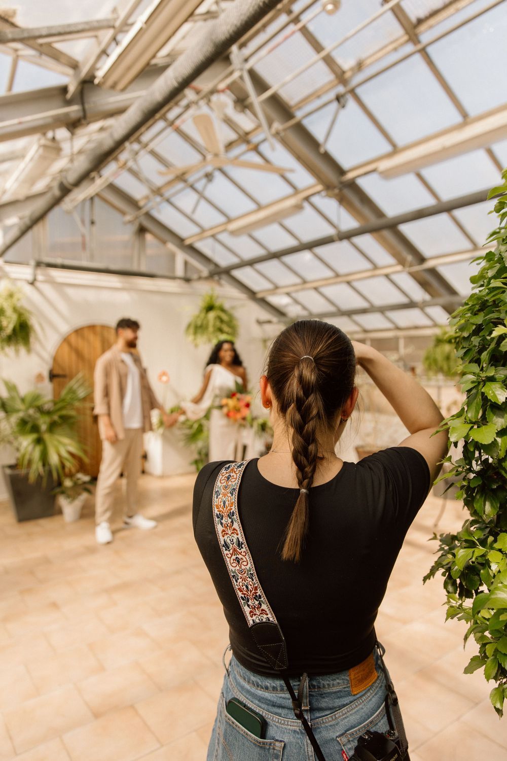 A photographer with a braided ponytail is taking pictures of a couple standing in a greenhouse. The couple is dressed in