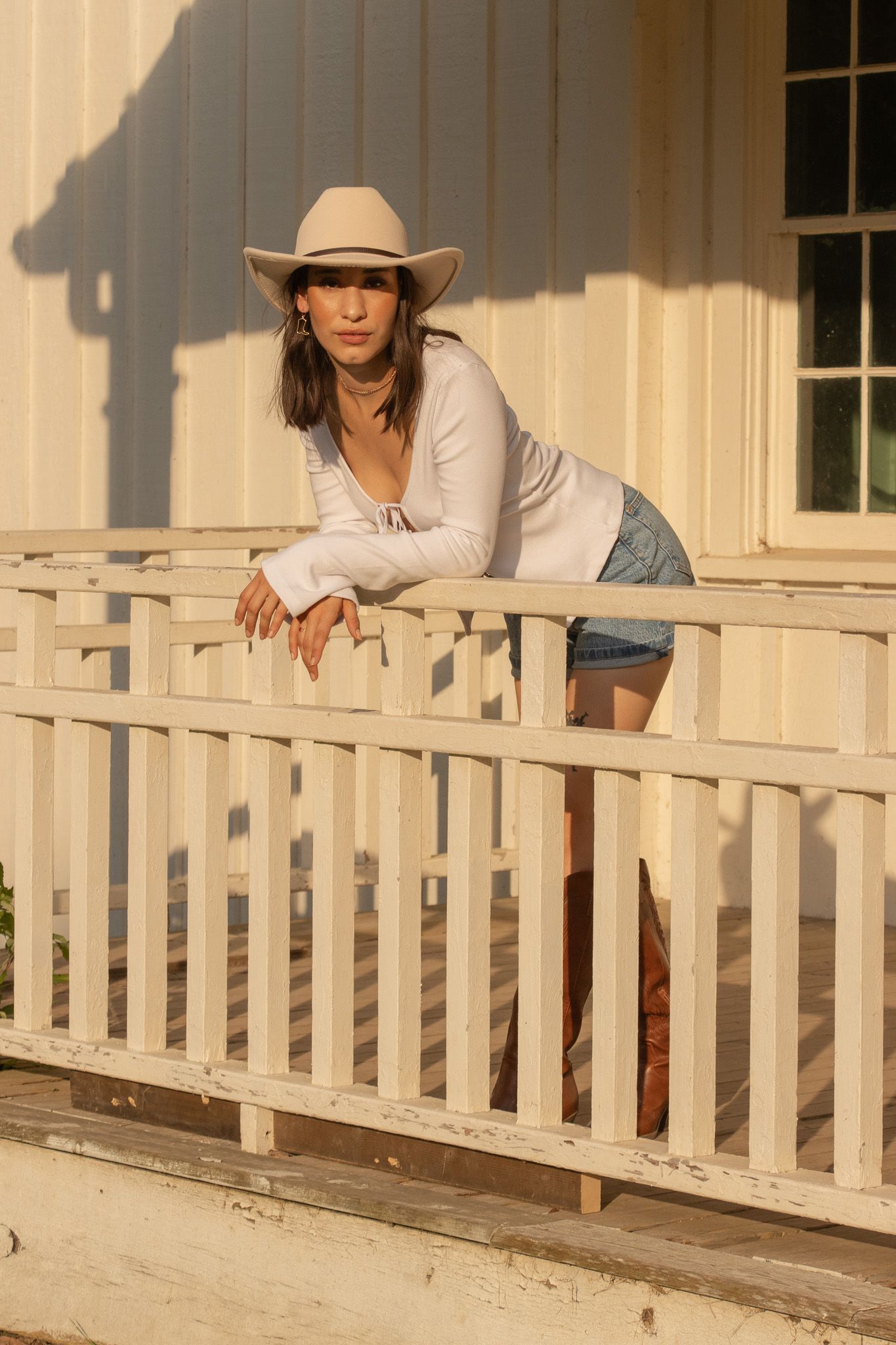 A woman wearing a white cowboy hat, white long-sleeve shirt, denim shorts, and brown cowboy boots leans on a wooden rail