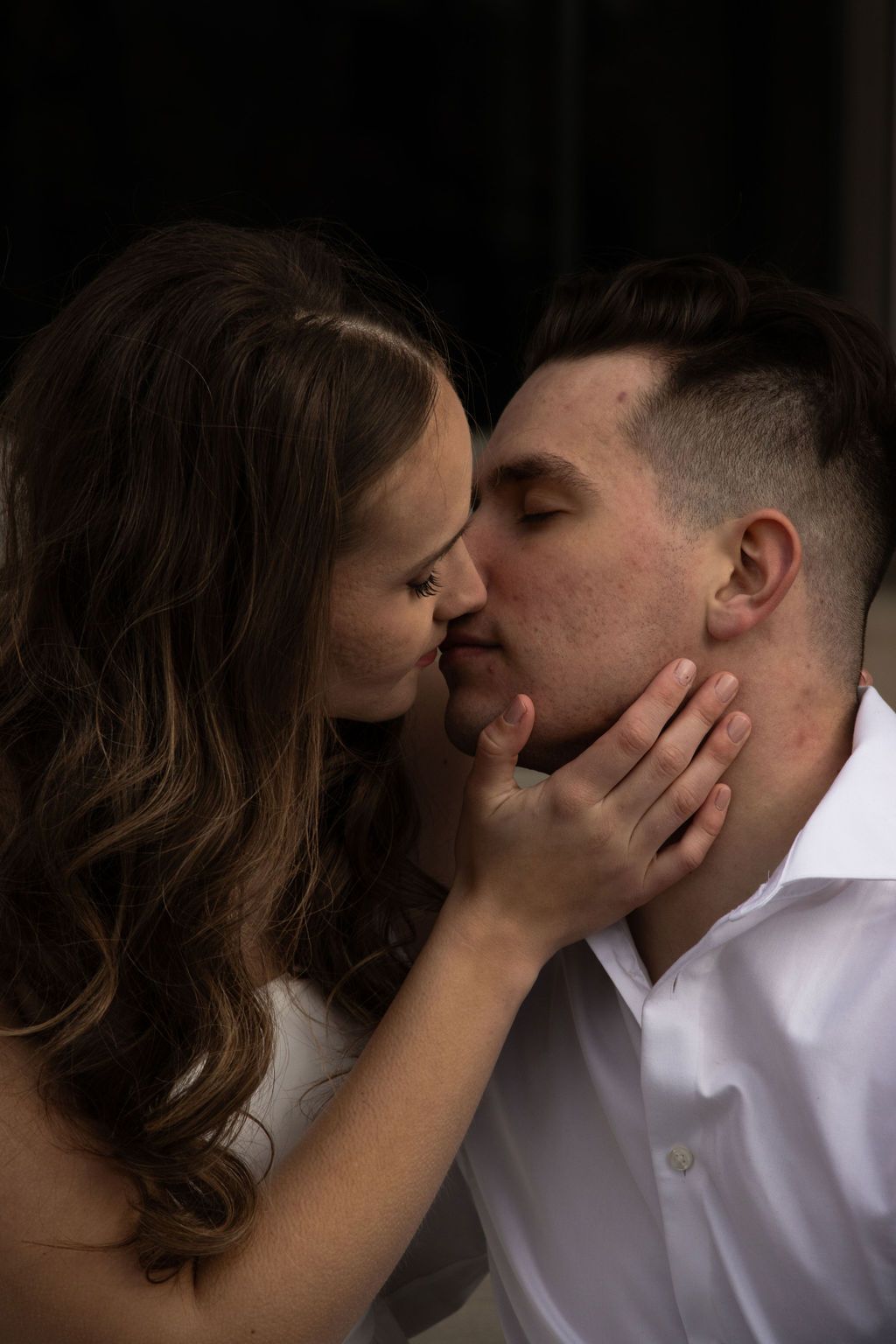 A woman with long, brown hair gently touches the face of a man with short, dark hair as they lean in for a kiss. Both ar