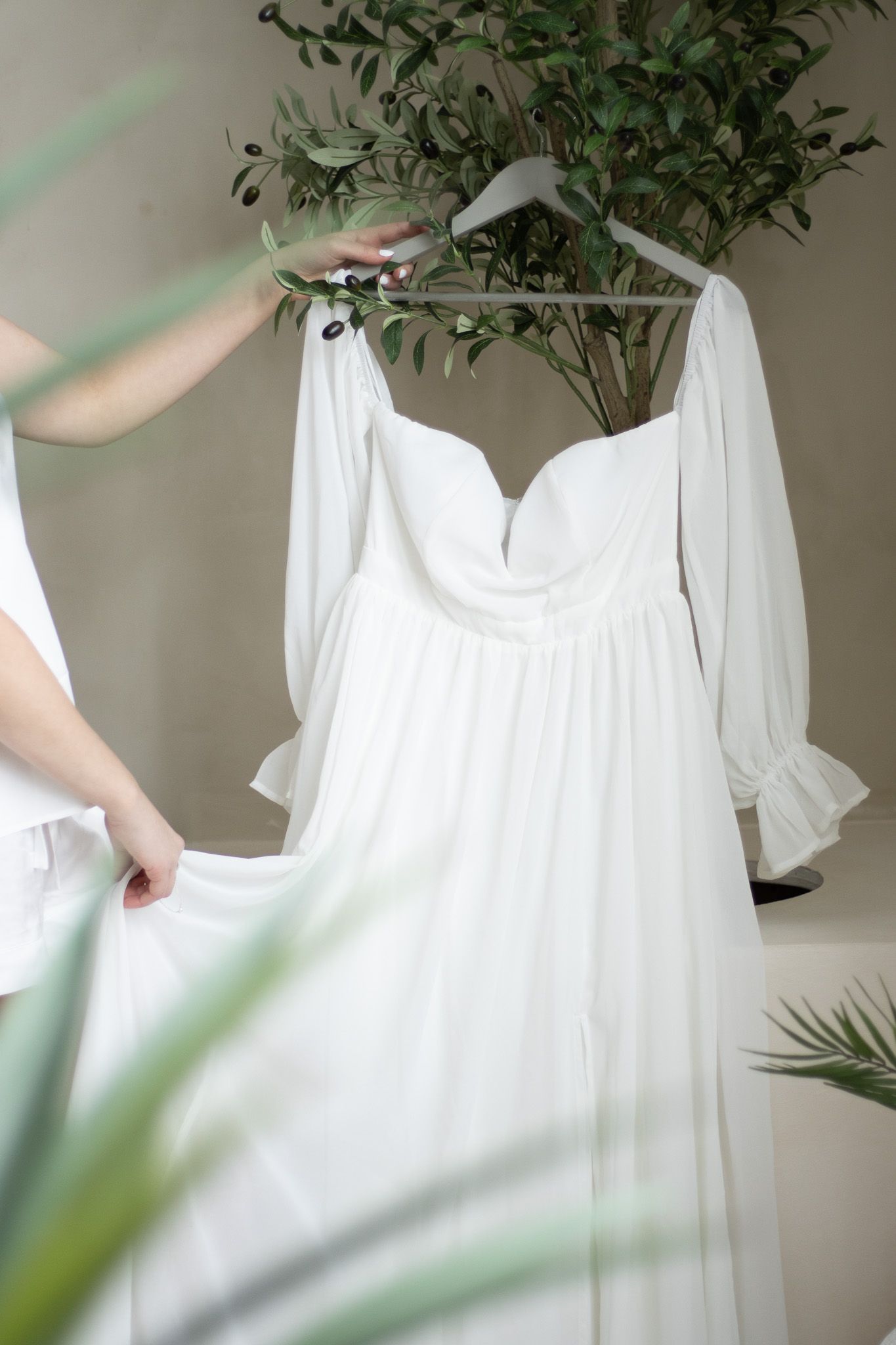 A person holding a white, long-sleeved, off-the-shoulder dress on a hanger, surrounded by green plants. The person is pa