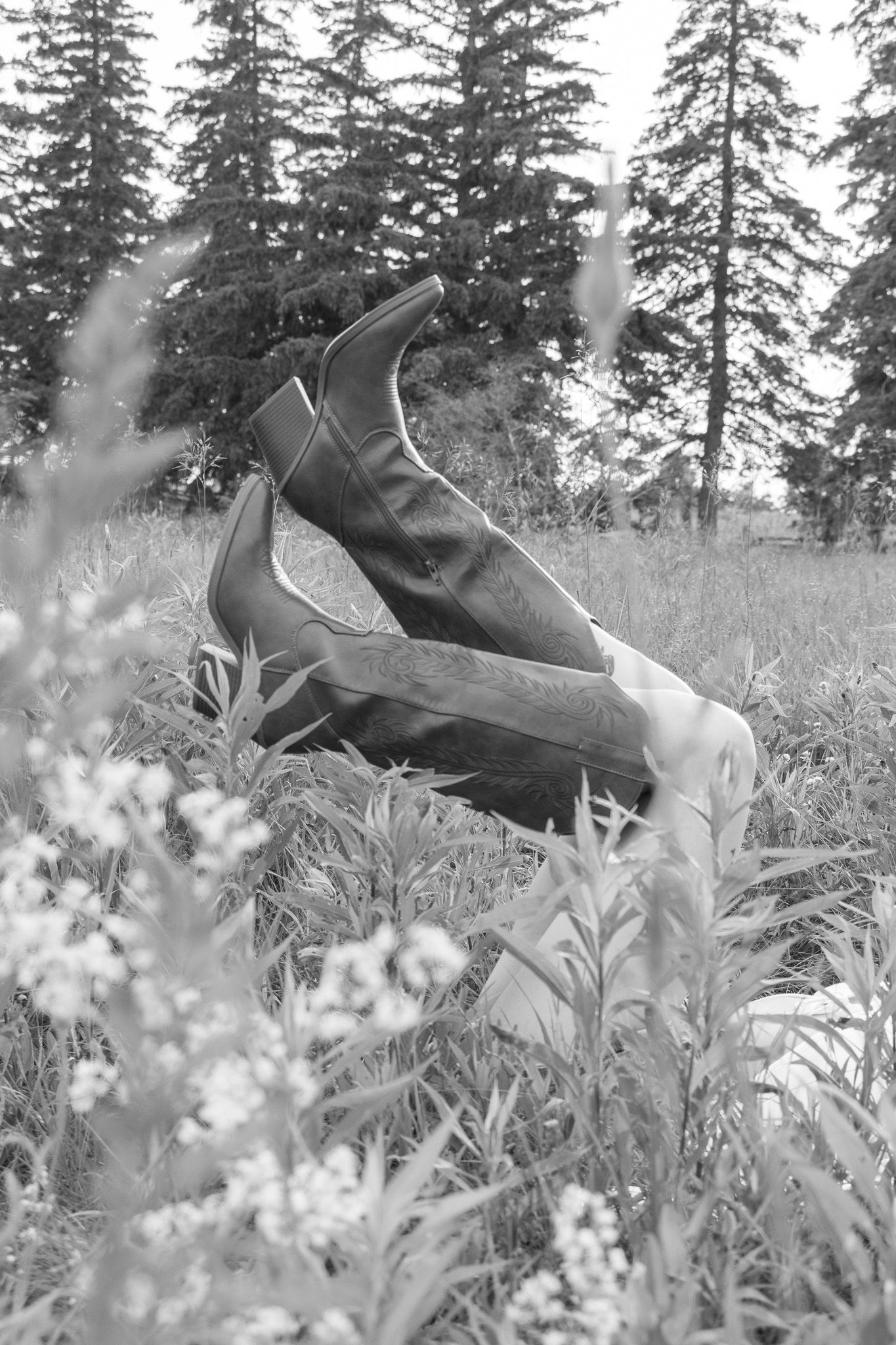 Black and white image showing a person's legs sticking straight up from a field of tall grass and wildflowers. The perso