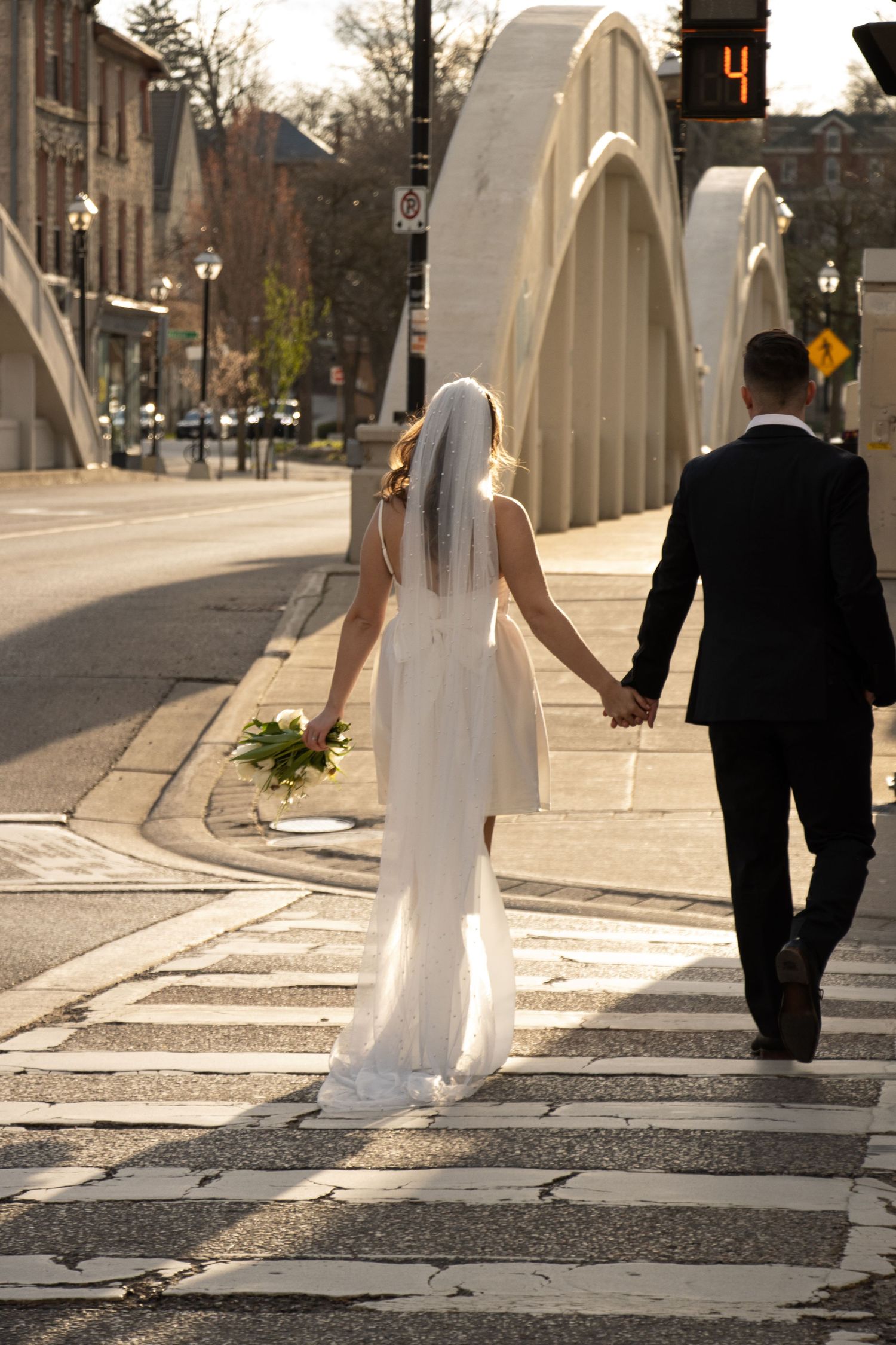 A bride in a white dress and veil holds a bouquet and walks hand in hand with a groom in a black suit across a crosswalk