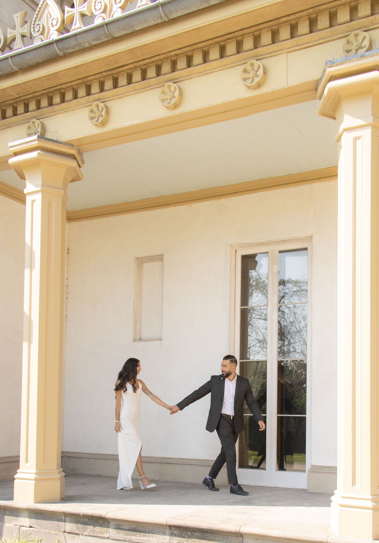 A couple, dressed elegantly, stand hand in hand on a porch with tall columns and decorative molding. The woman wears a l