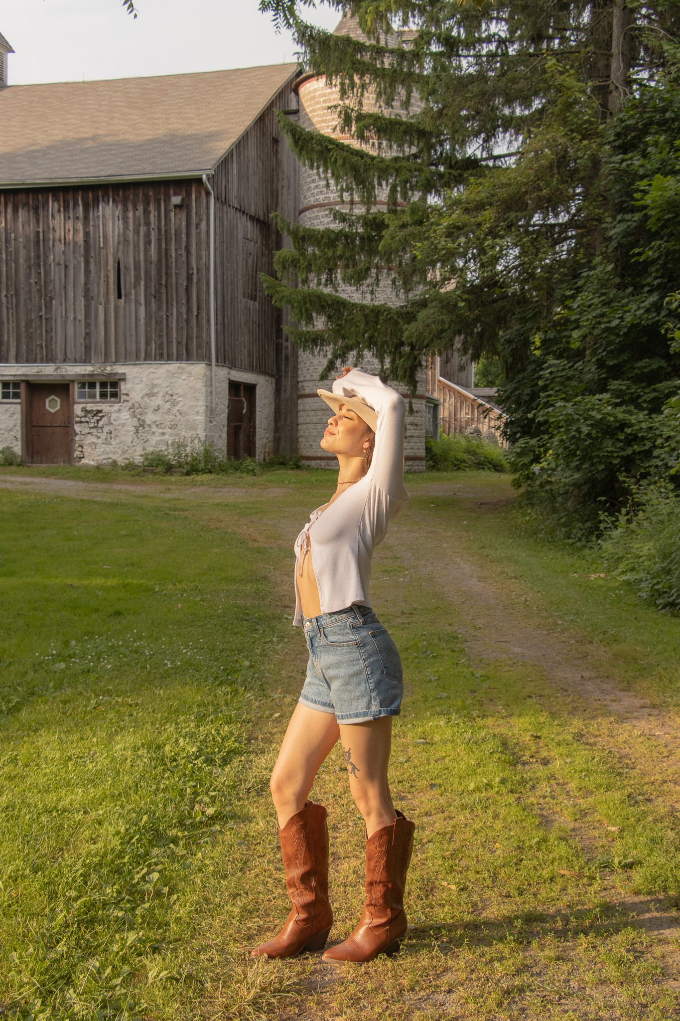 A person wearing a long-sleeve top, denim shorts, and cowboy boots stands on a grassy path with one hand shielding their