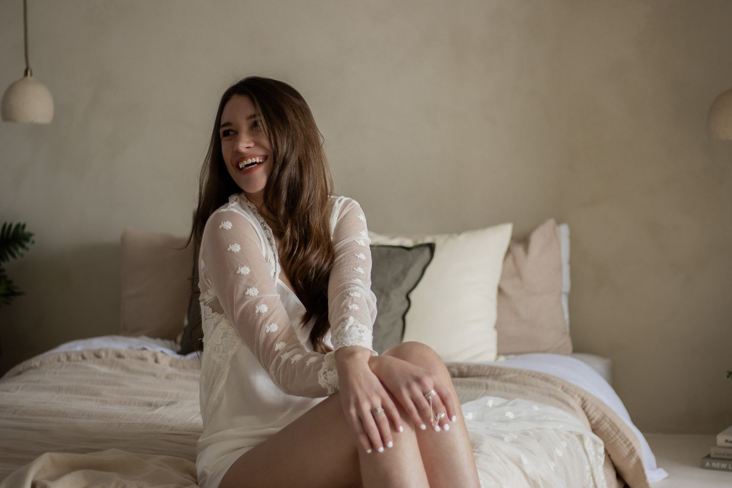 A woman sitting on the corner of the bed, with her hands resting on their knees. The person is wearing a lace-covered ro