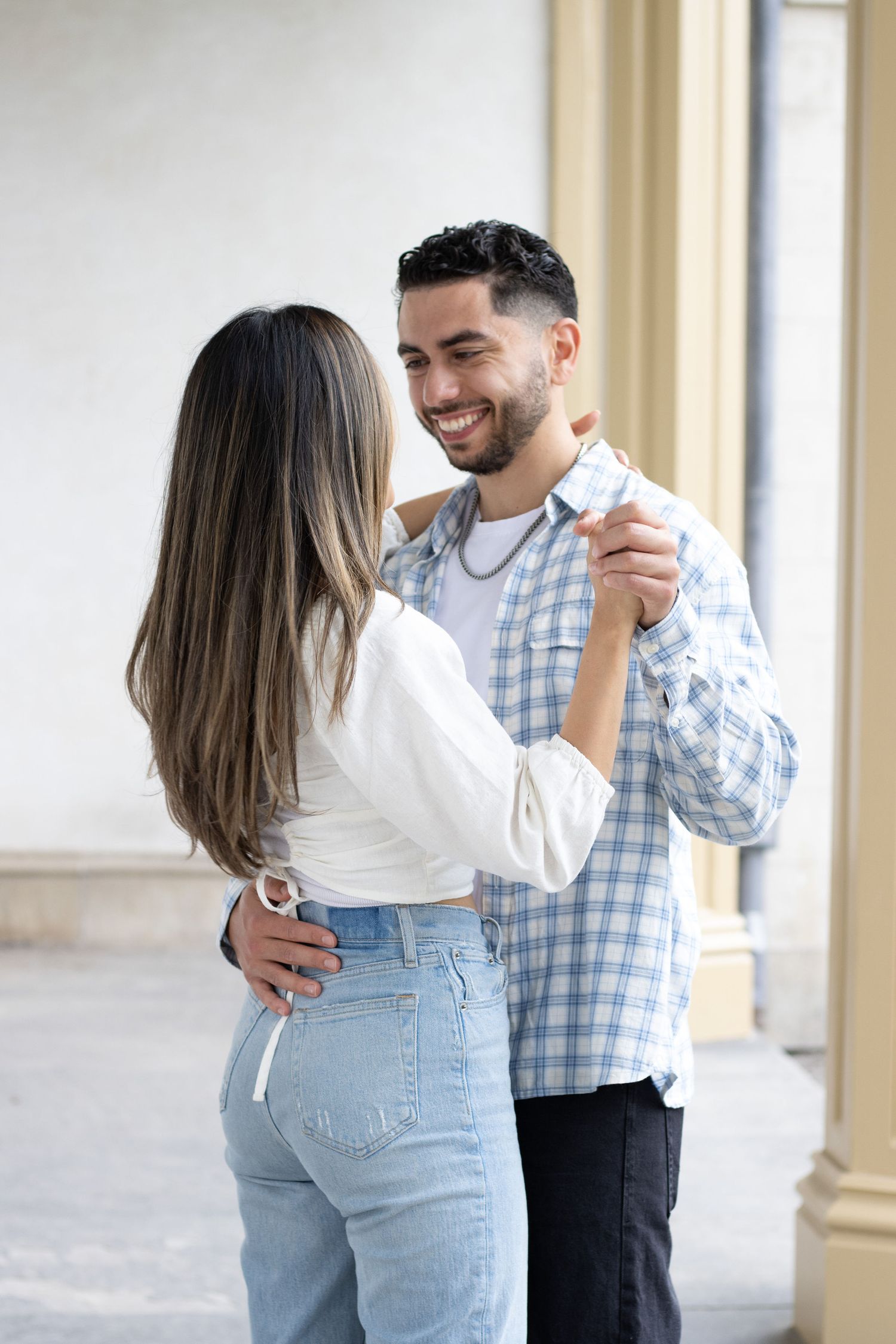A couple slow dancing, in an outdoor corridor. The woman is wearing a white long-sleeve top and blue jeans, while the ma