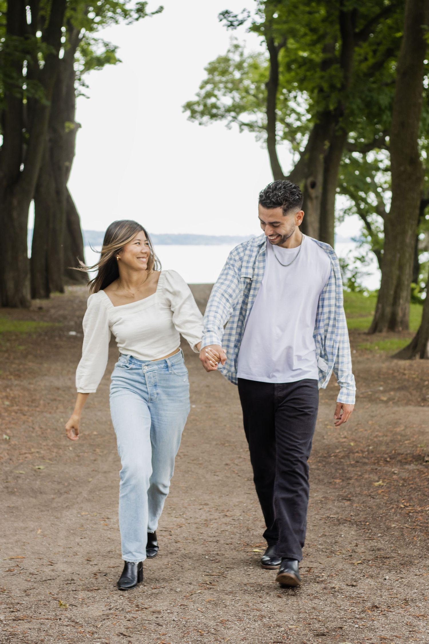 A couple holding hands walks together in a wooded path. The woman is wearing a white long-sleeve top and blue jeans, whi