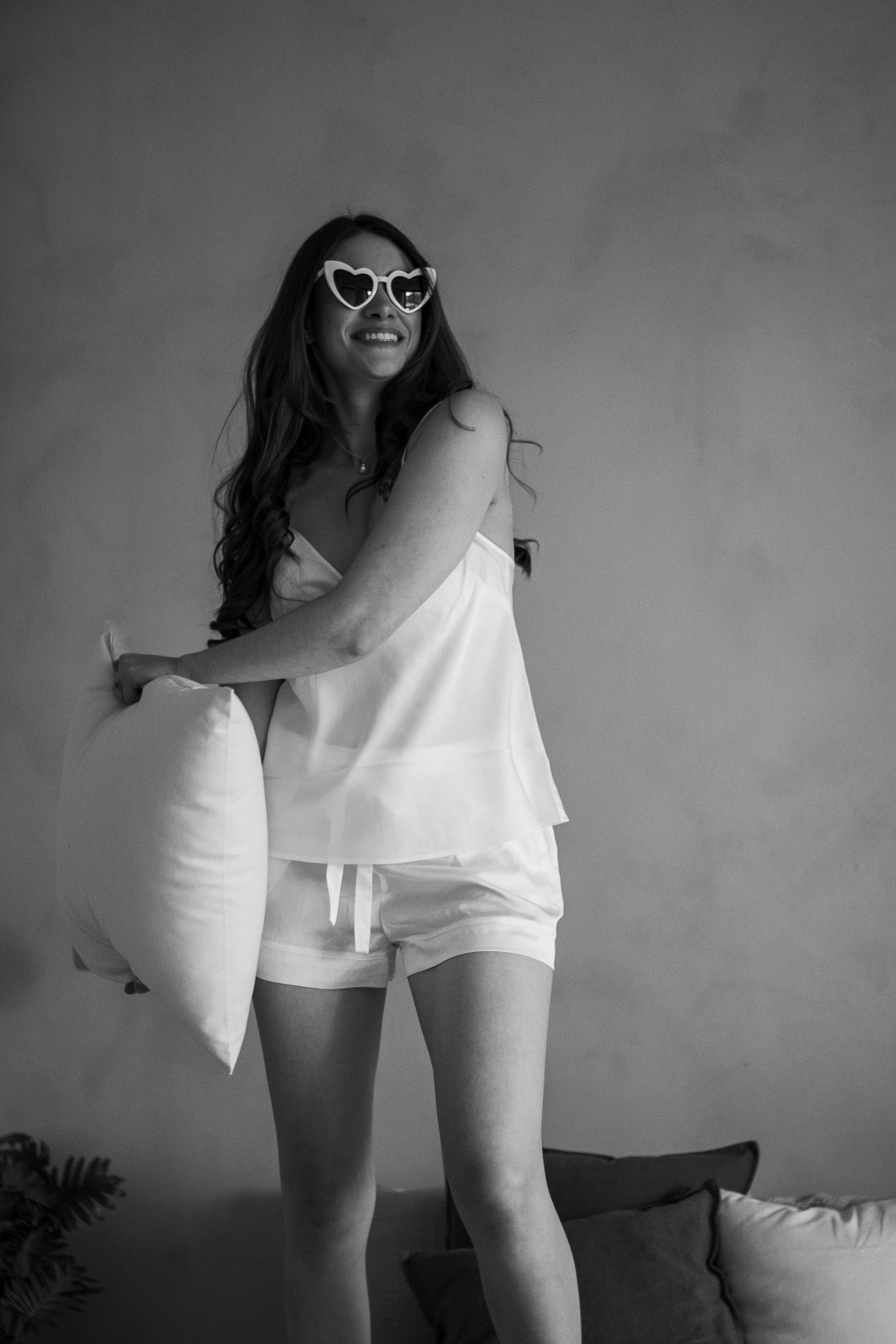 A woman dressed in white sleeveless top and shorts is holding a pillow. She is smiling and wearing heart-shaped sunglass