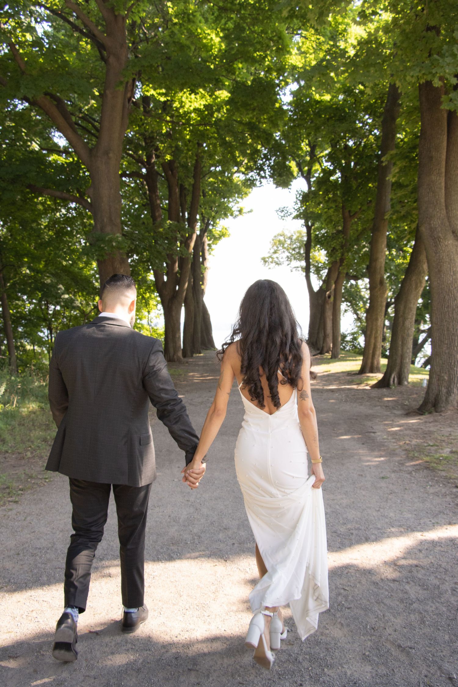 A couple dressed in formal attire, with the woman in a white gown and the man in a black suit, walk hand-in-hand down a 
