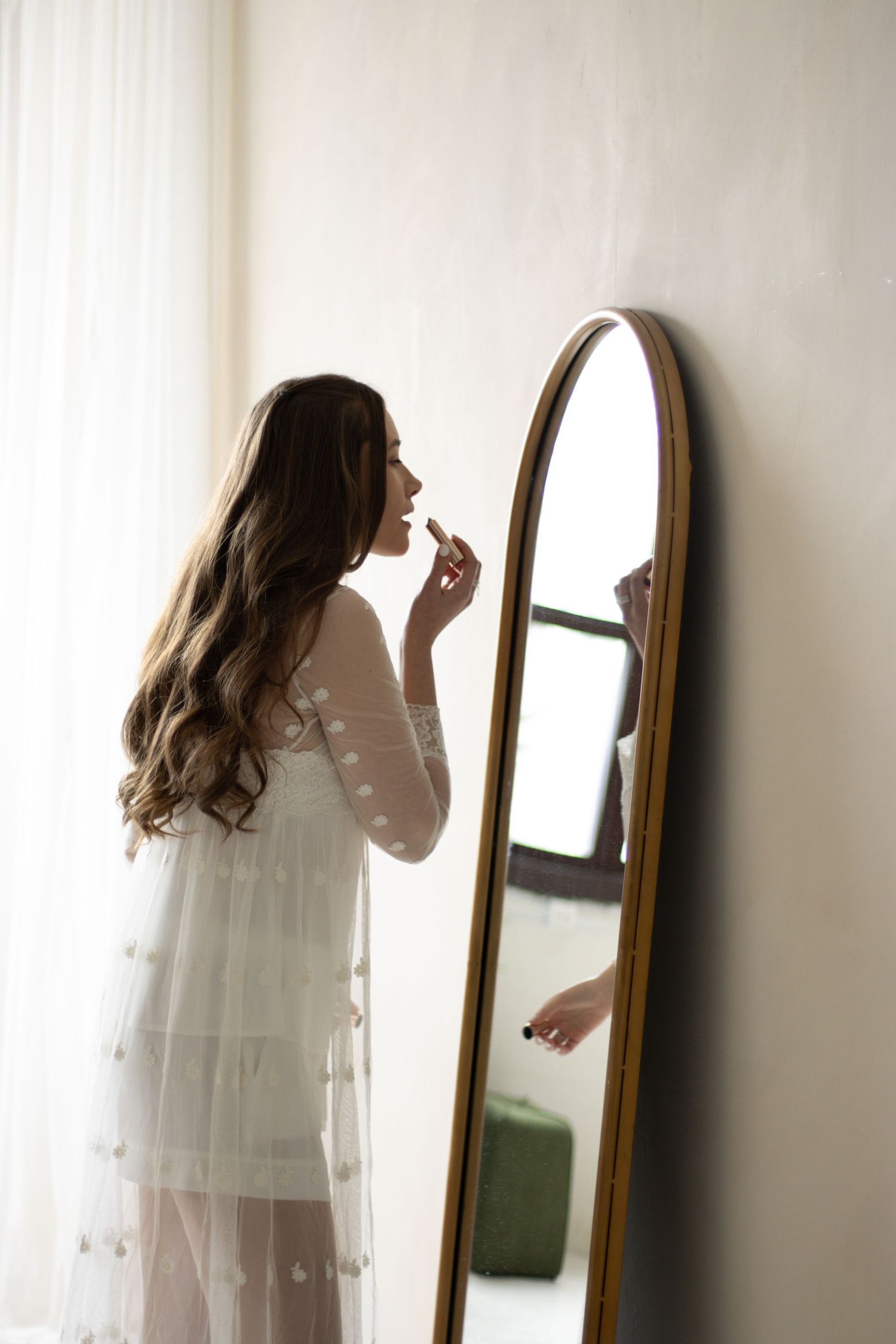 A woman with long, wavy hair and wearing a white, sheer dress is applying lipstick in front of a tall, oval mirror. The 