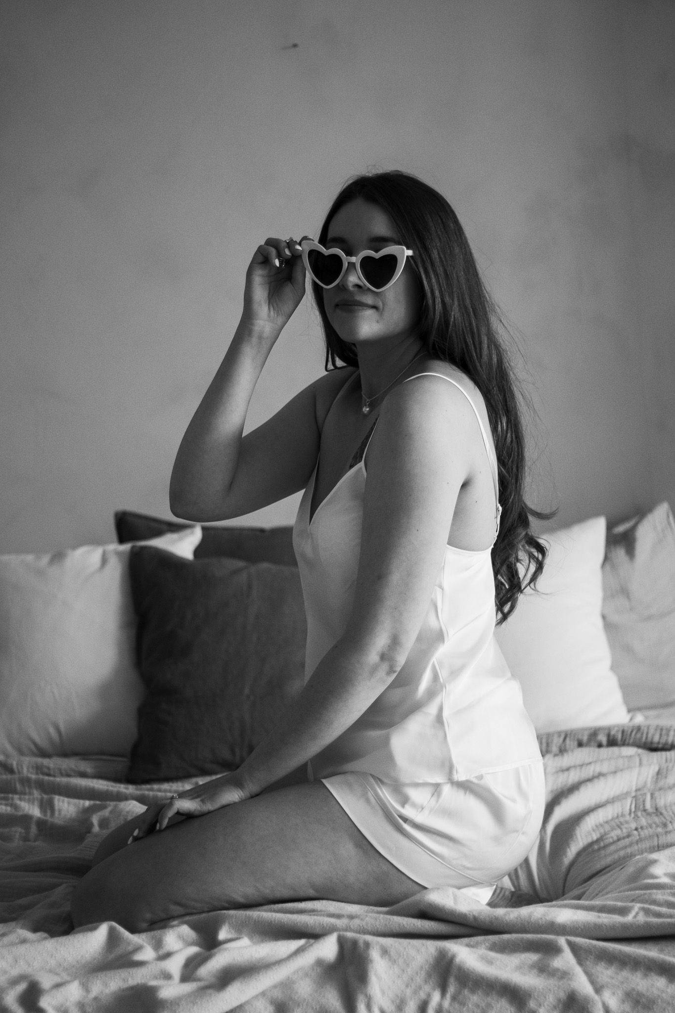 A woman dressed in white sleeveless top and shorts is kneeling on a bed. She is looking at the camera and holding her he