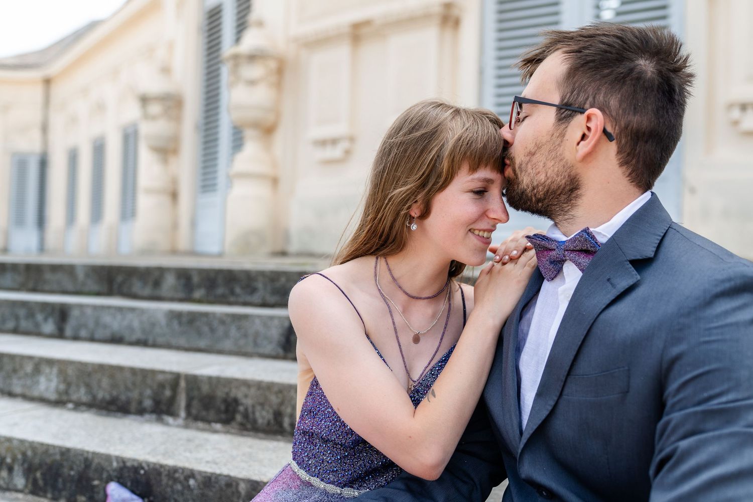 A tender moment between a couple on stone steps with her in a purple dress and him in a bowtie.