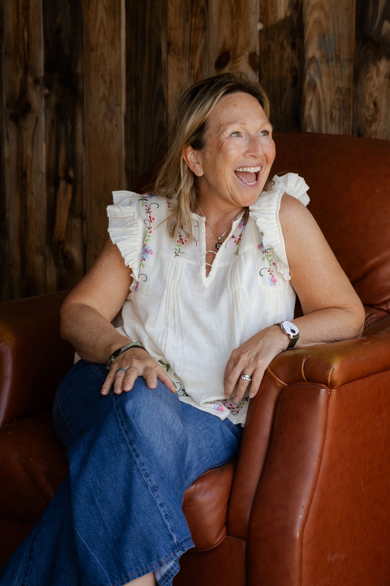A smiling woman in a white floral blouse and jeans sits laughing in a brown leather chair against a rustic wood wall.