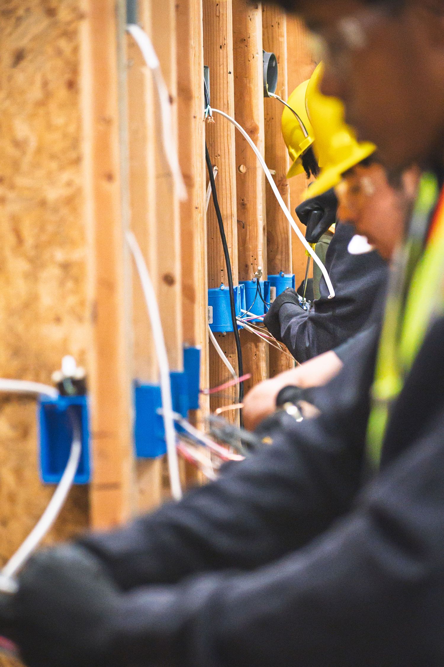 Electricians in yellow hard hats installing wiring in blue electrical boxes on wooden wall framing at a construction site.