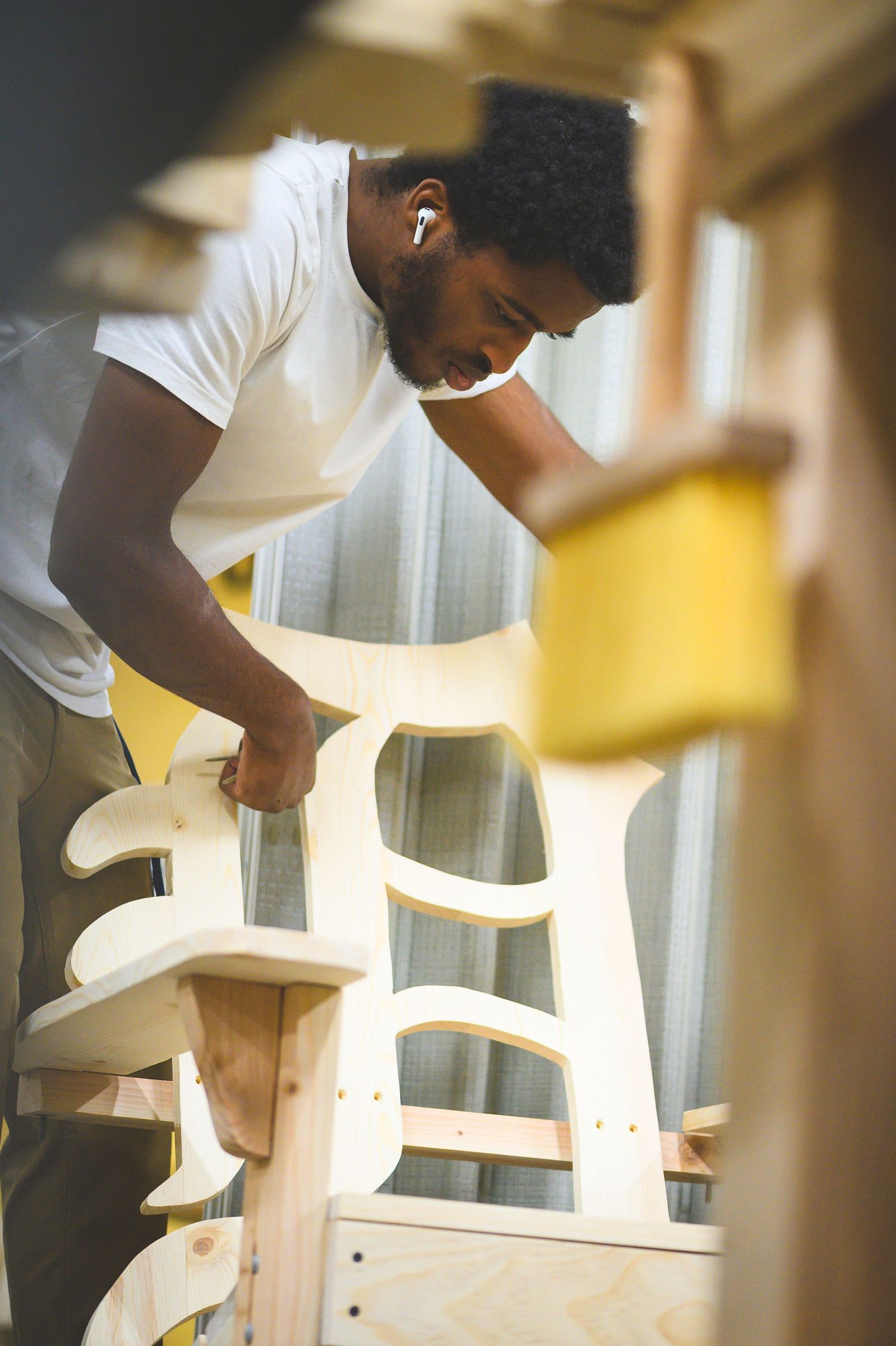 A person in a white t-shirt assembles a white wooden chair near a window with natural light streaming in.