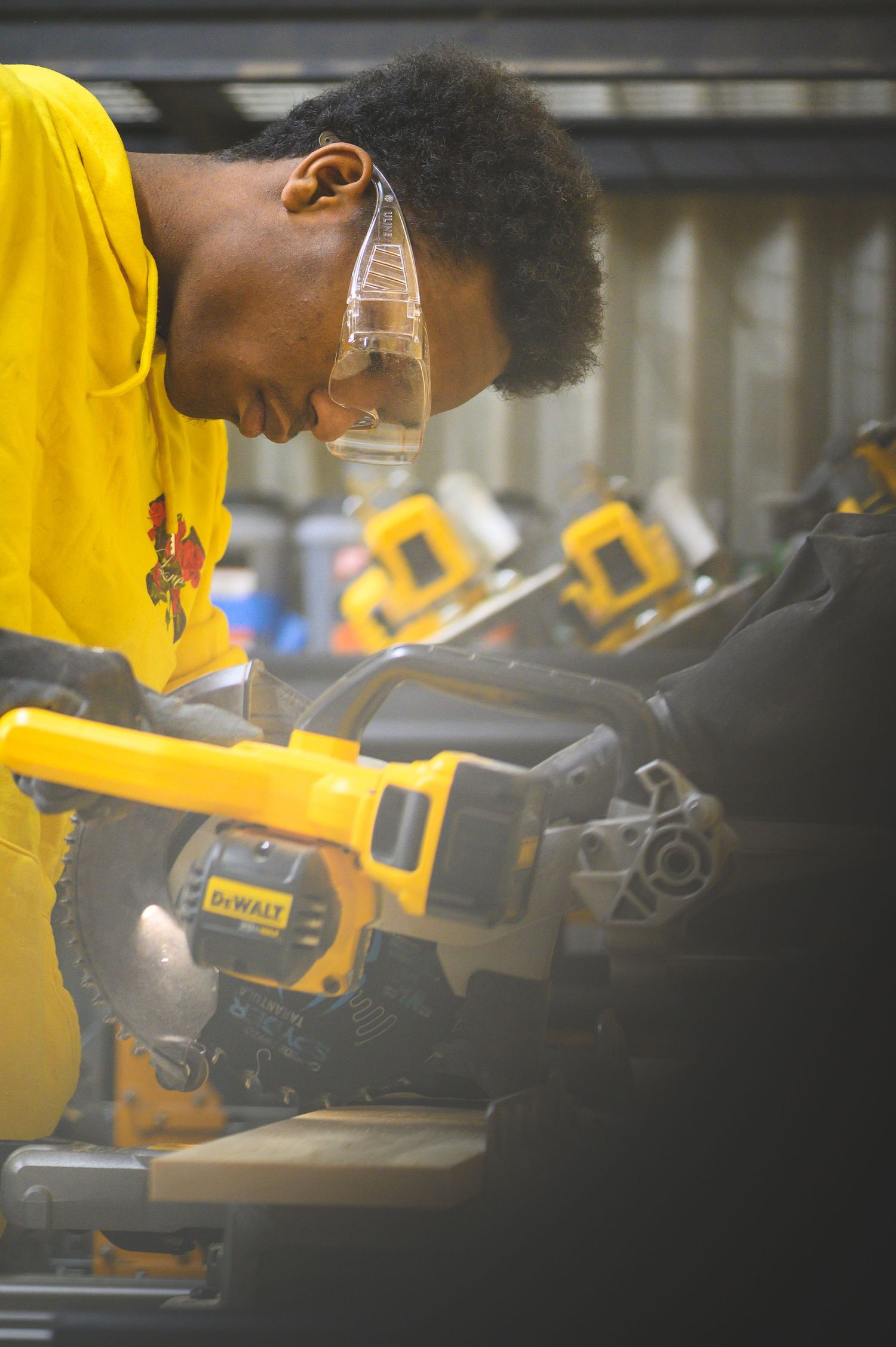 Student in safety glasses and yellow shirt carefully operates DeWalt power tools in workshop setting.