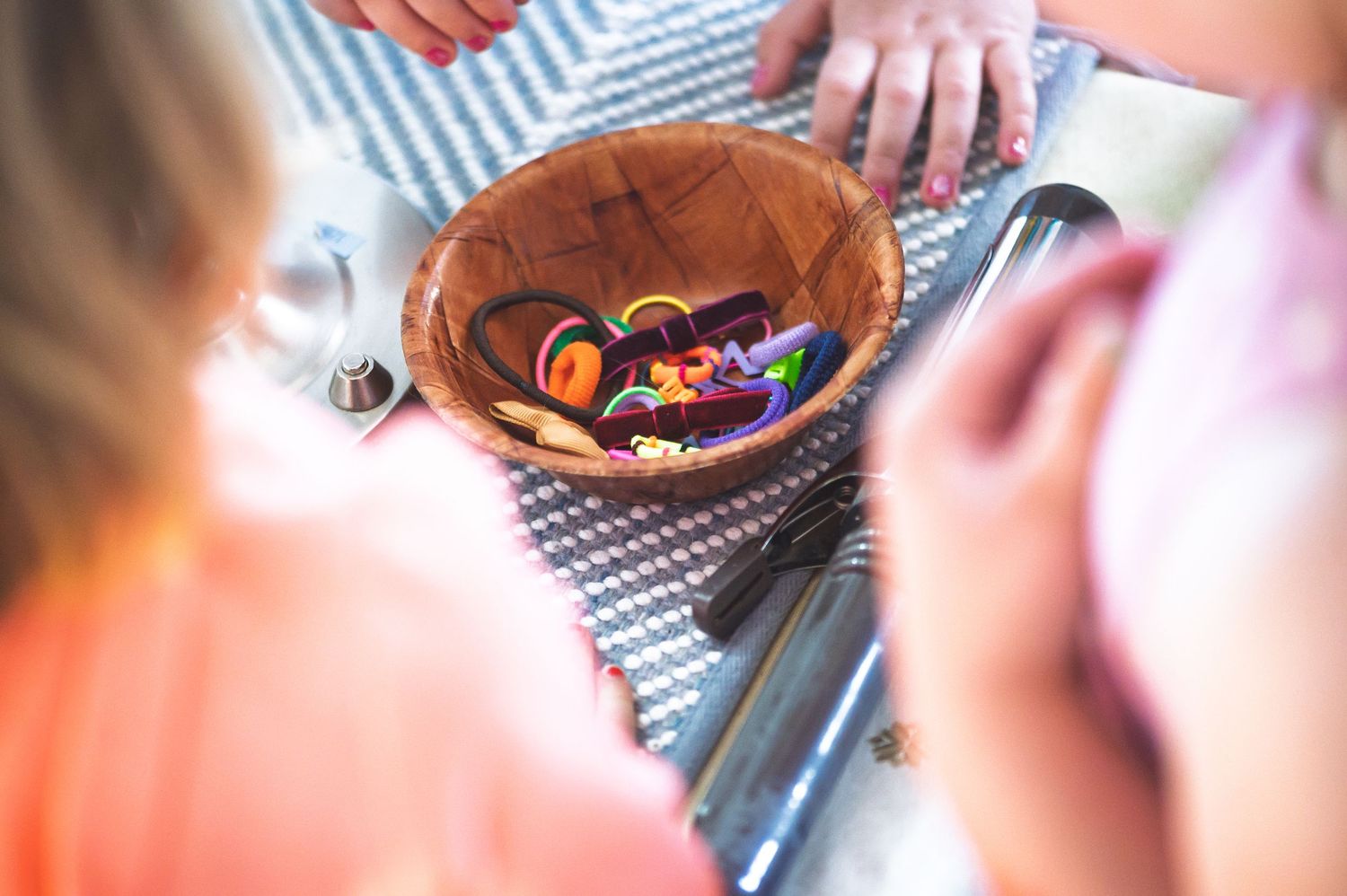 Children gathered around a wooden bowl filled with colorful hair accessories on a striped mat.