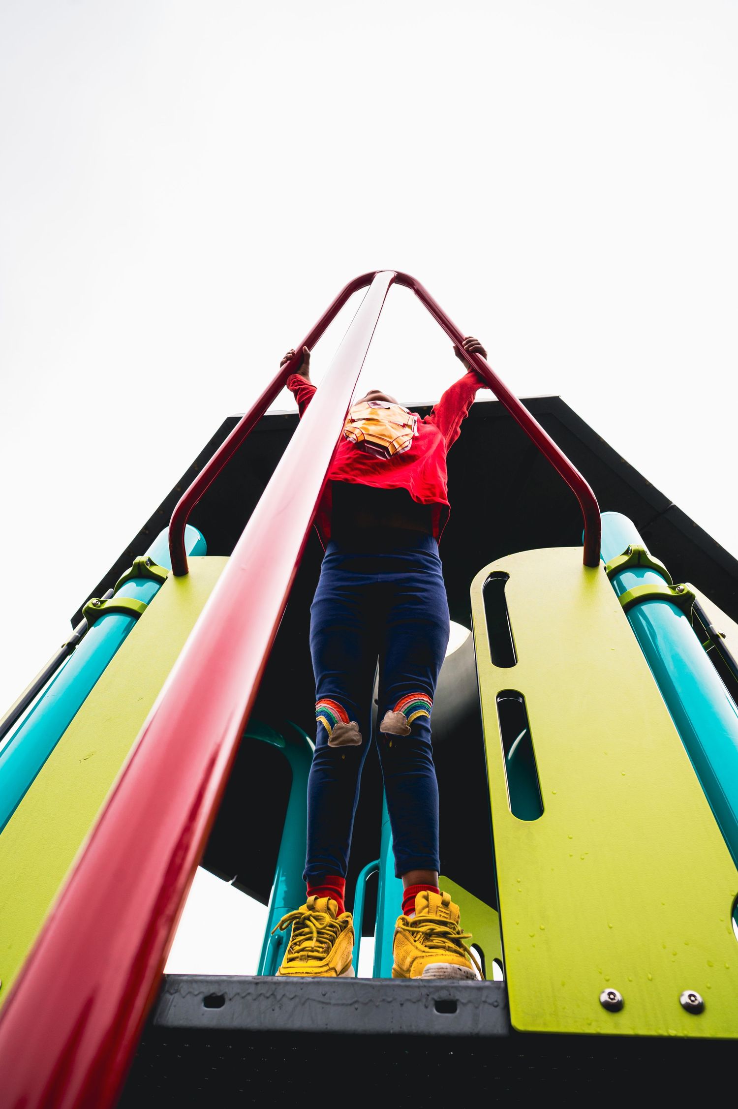 Child in red sweater and yellow sneakers climbing colorful playground equipment viewed from below against white sky.