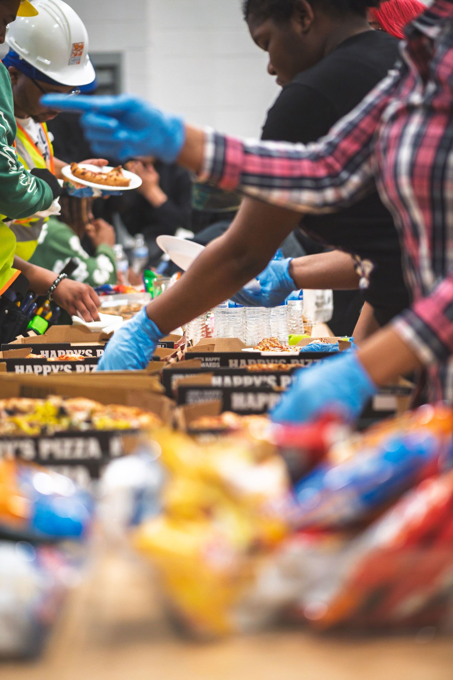 Volunteers wearing blue gloves serve pizza and food at a community charity event with Happy's Pizza boxes on the table.