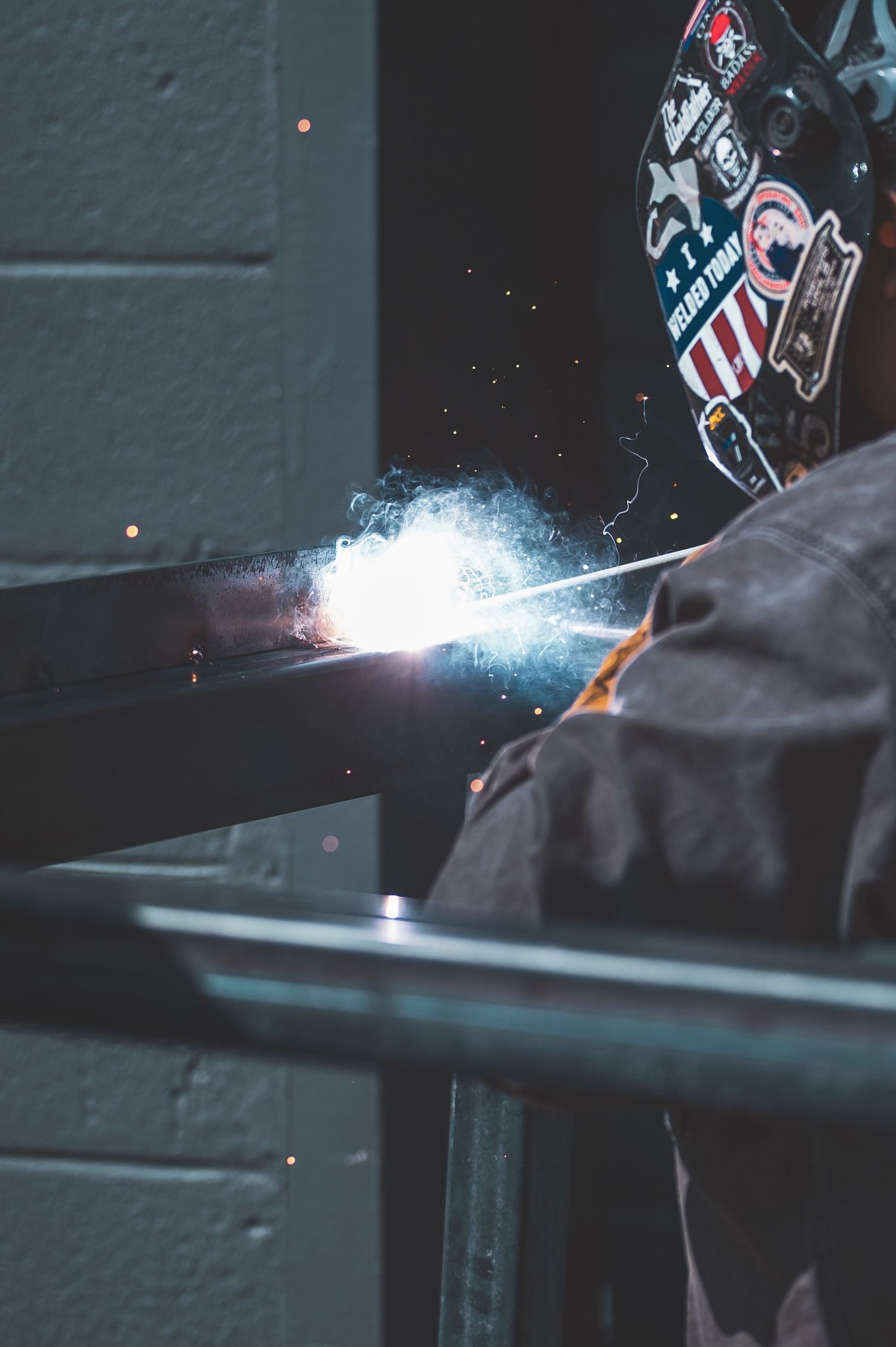 A welder wearing a sticker-covered helmet creates bright sparks while welding metal beams.