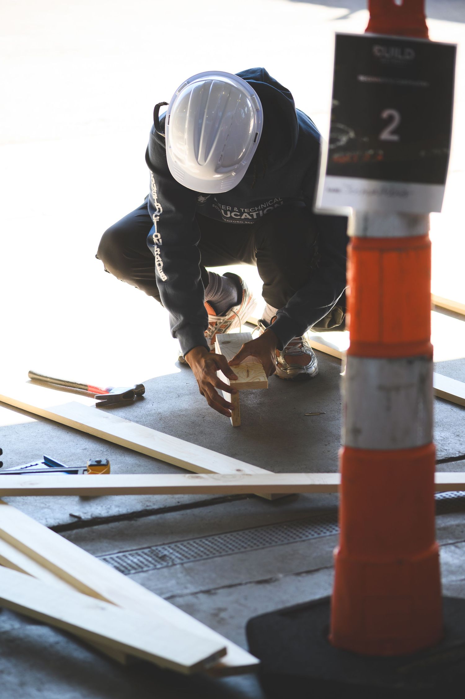 Construction worker in hard hat crouching over wooden planks near an orange traffic cone at a worksite.