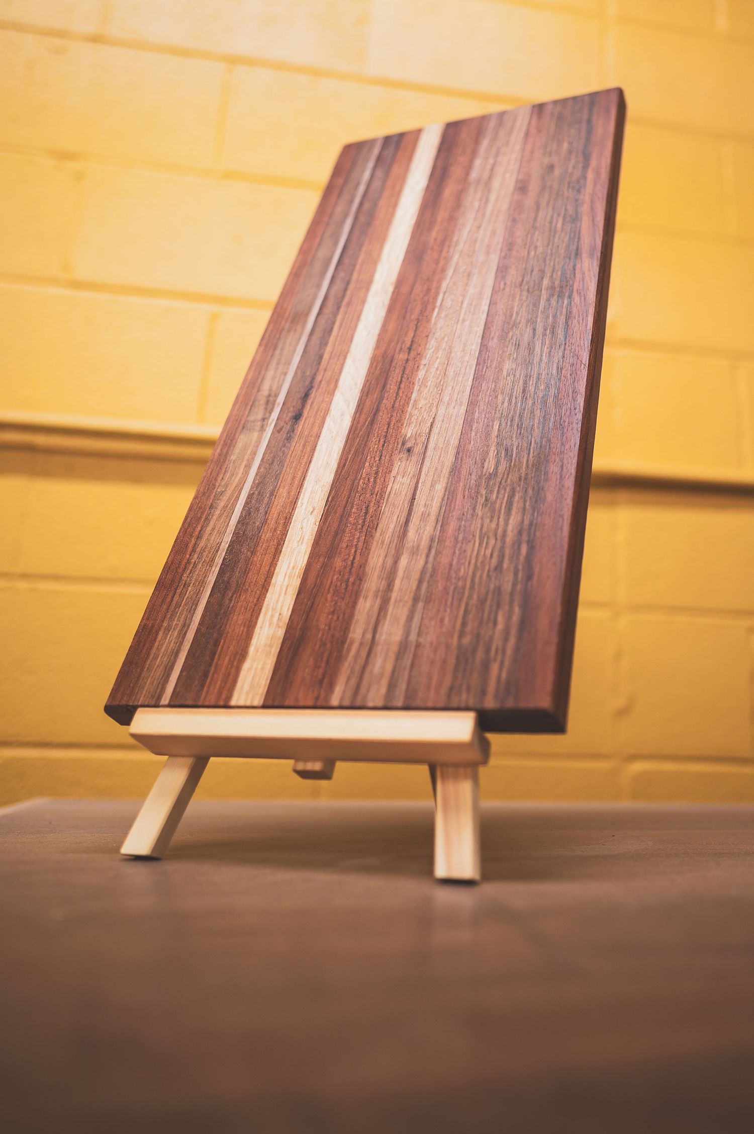 Handcrafted wooden cutting board with walnut and maple stripes displayed on a small easel stand against a yellow brick wall.