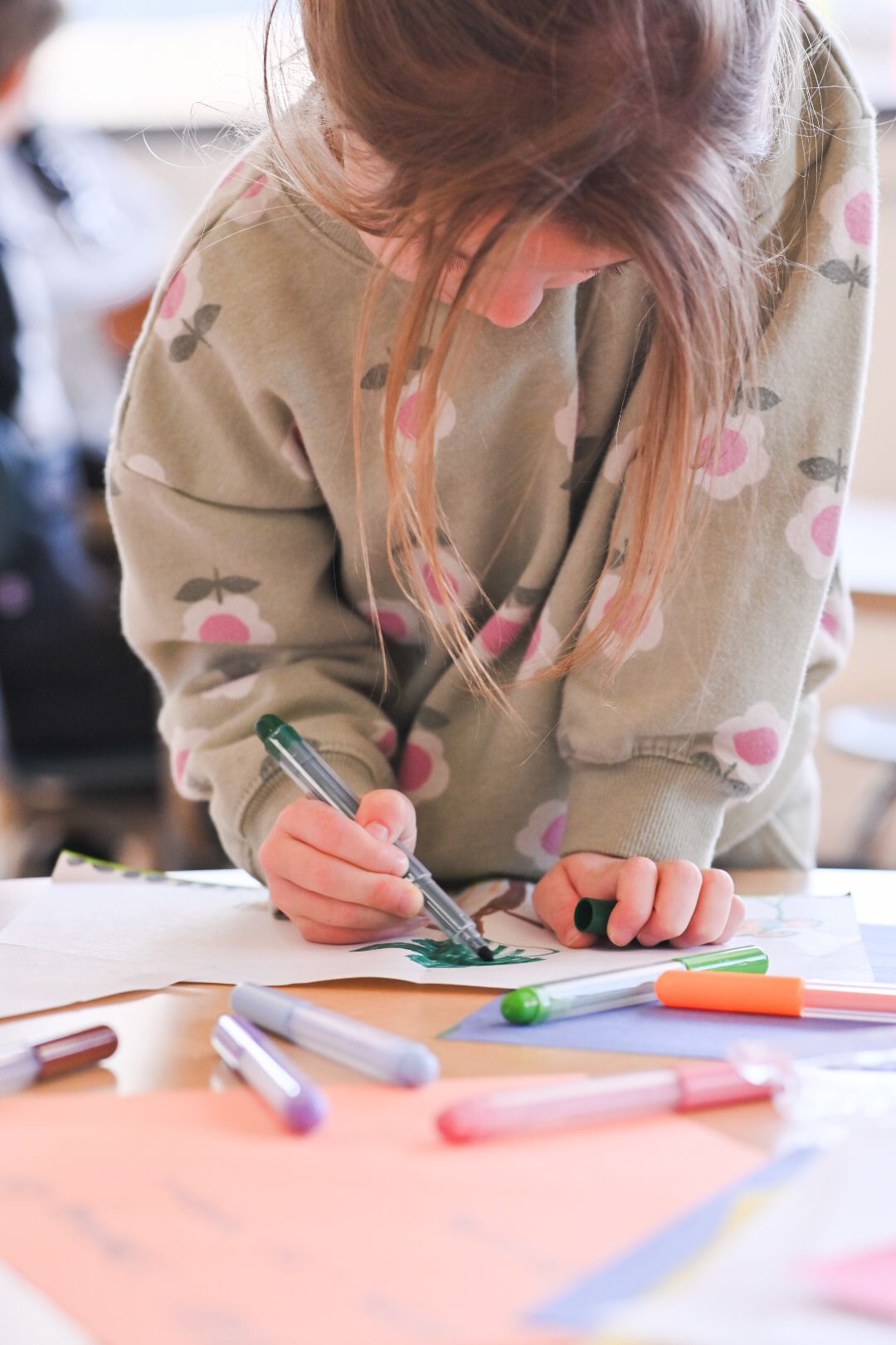 Young girl drawing with green marker on white paper surrounded by colorful markers on a table.