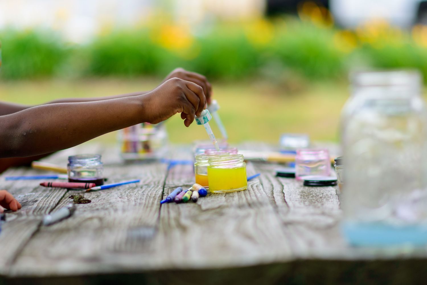 Child dipping a paintbrush into yellow paint jar on a rustic wooden table during an outdoor art activity.