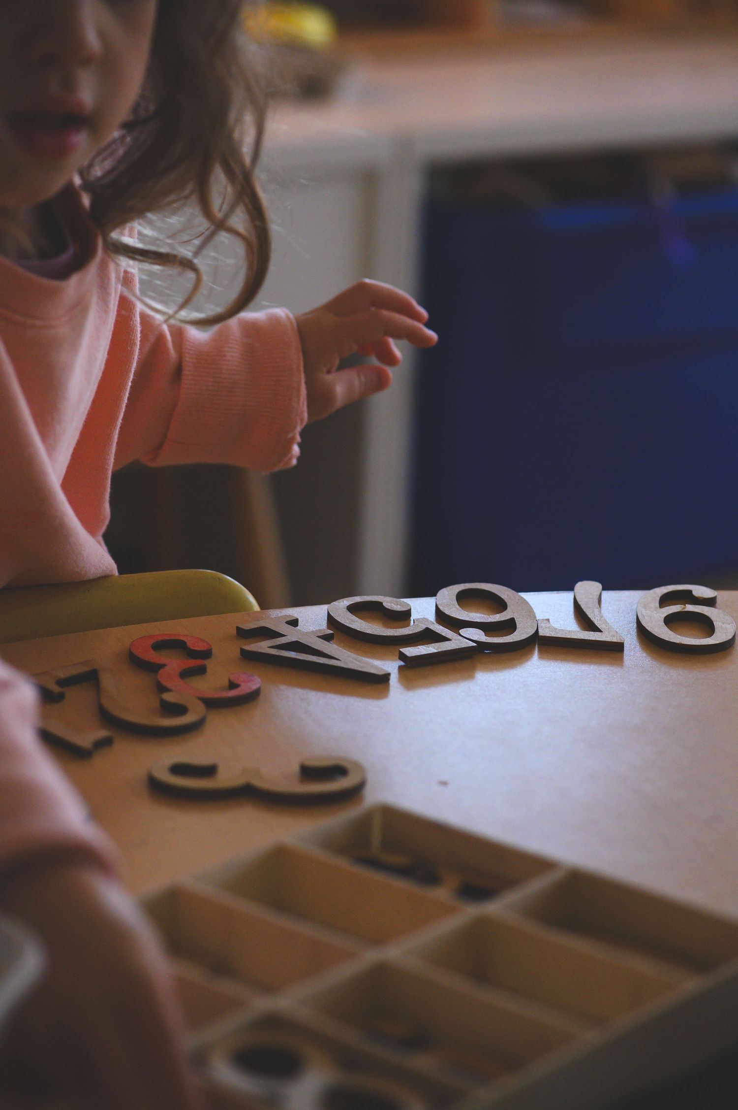 Young girl in pink sweater playing with wooden number and letter puzzle pieces on a table.