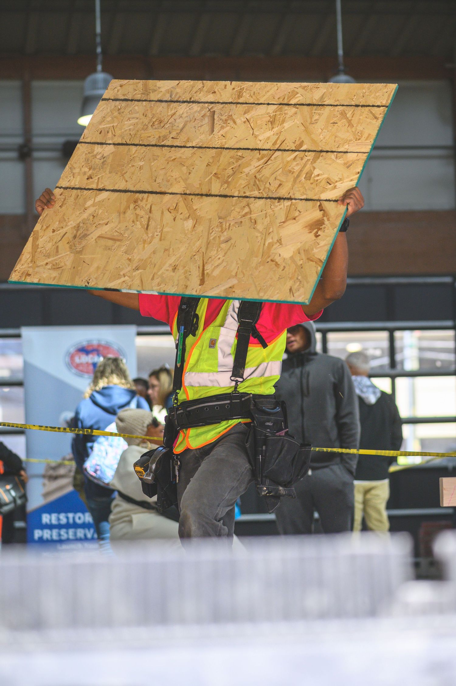 Construction worker in safety vest carries large plywood sheet at an indoor worksite with yellow caution tape visible.