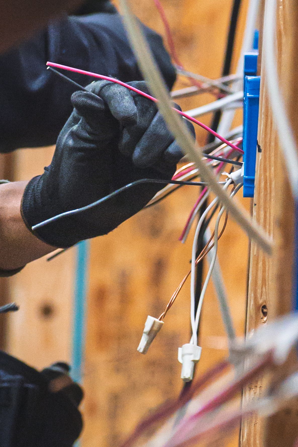 Electrician in black gloves working with colorful wires on a wooden wall electrical box during installation.