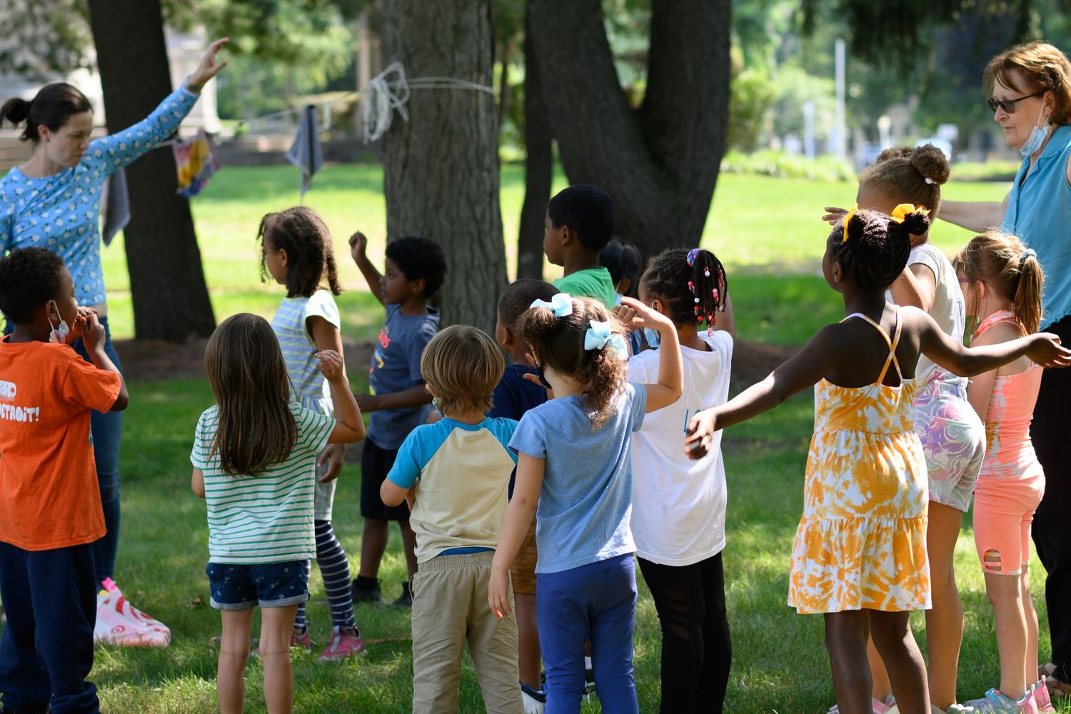 Children playing outdoor games in a sunny park guided by two adult supervisors on a warm summer day.