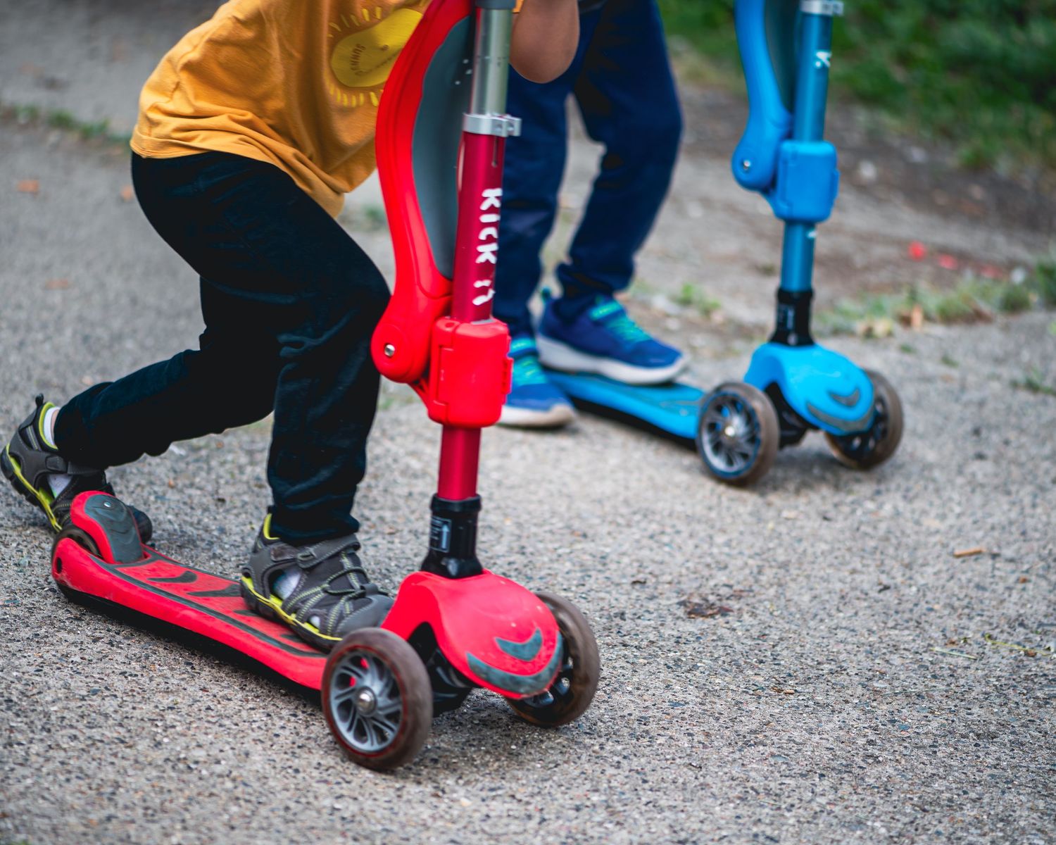 Two children riding colorful red and blue three-wheeled kick scooters on a paved path outdoors.