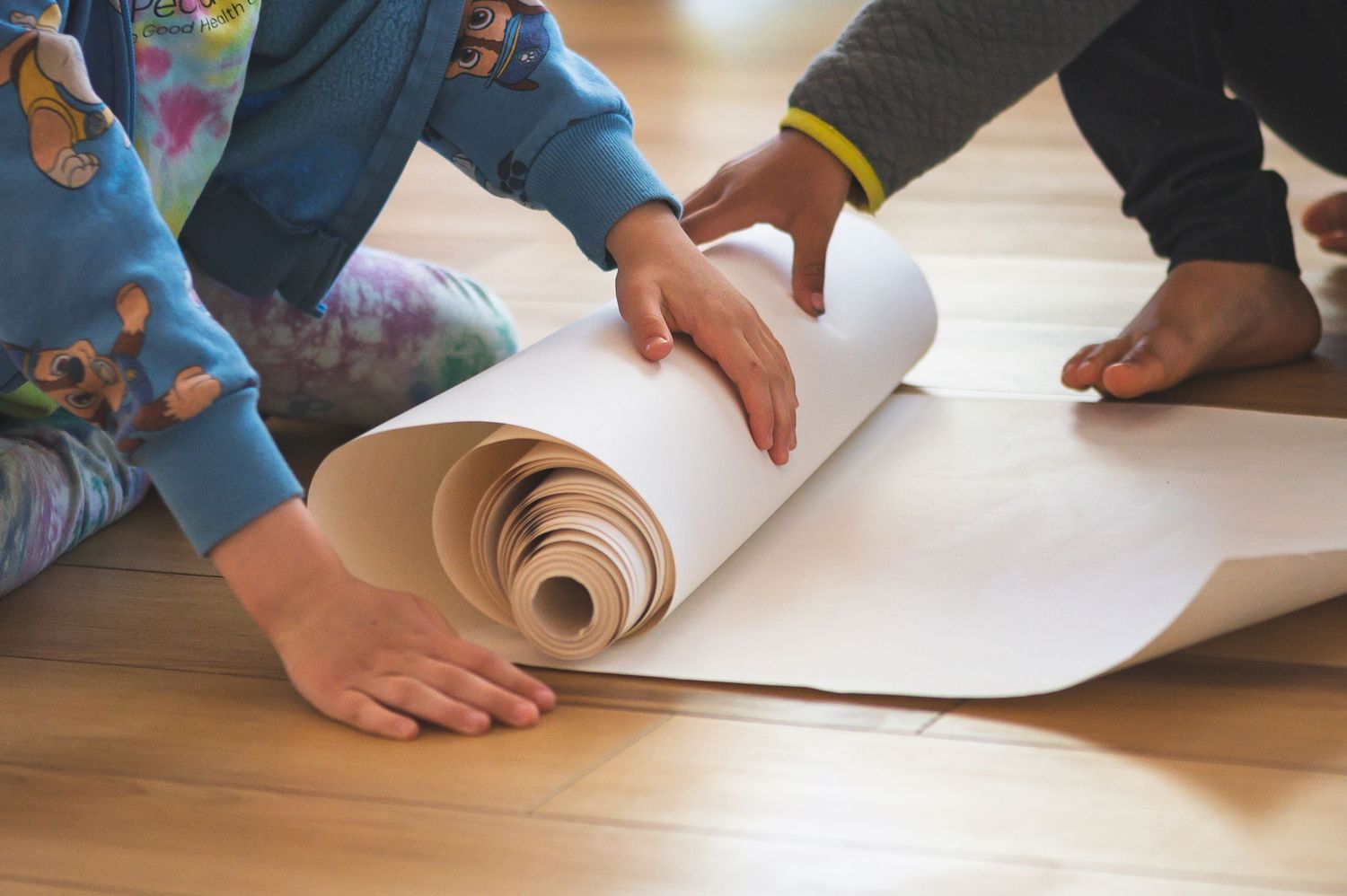Two children unrolling a large sheet of white paper on a wooden floor for an art project.