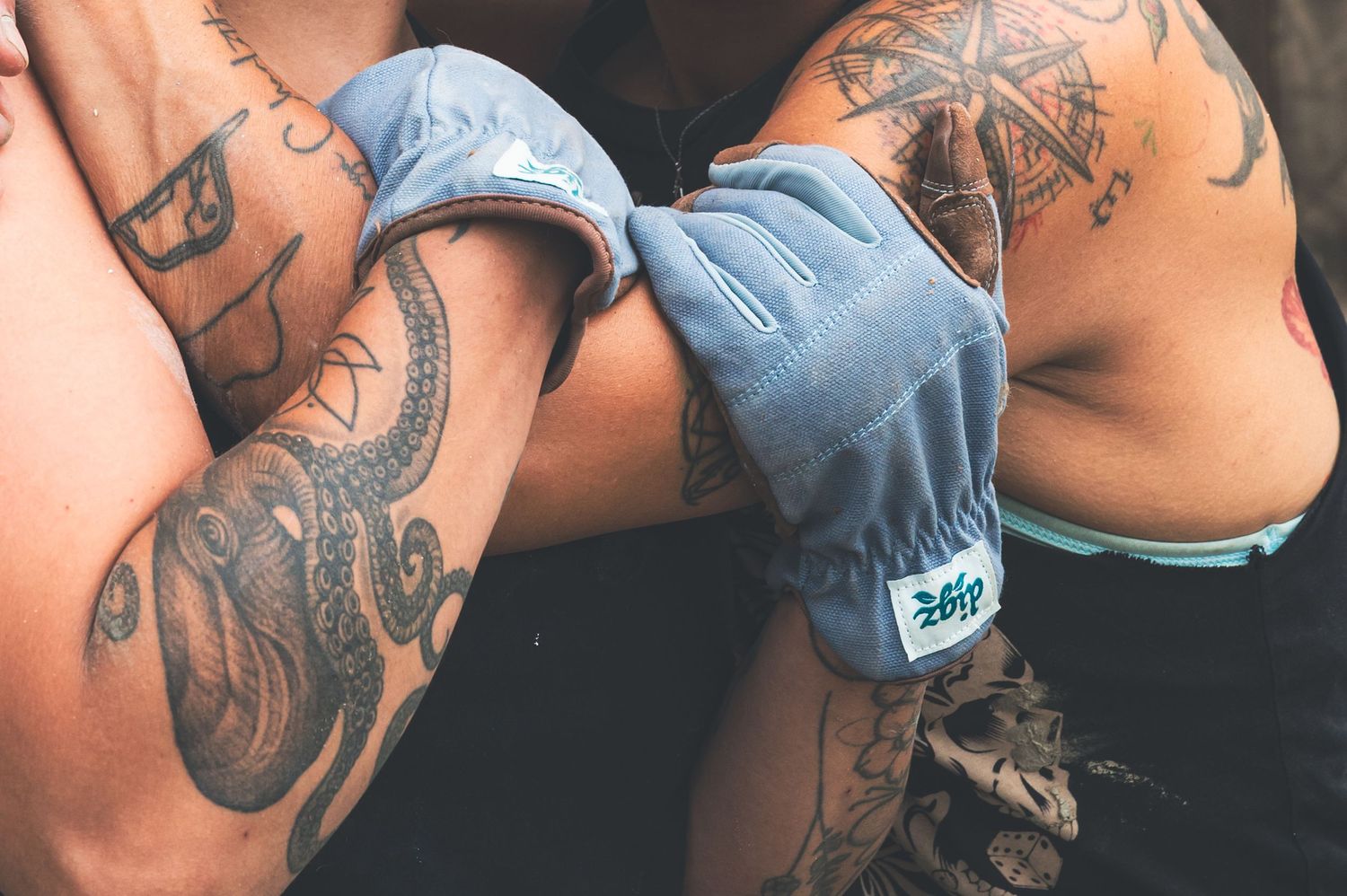 Close-up of tattooed arms wearing blue fingerless gloves with a brand logo, showcasing detailed tattoo artwork.