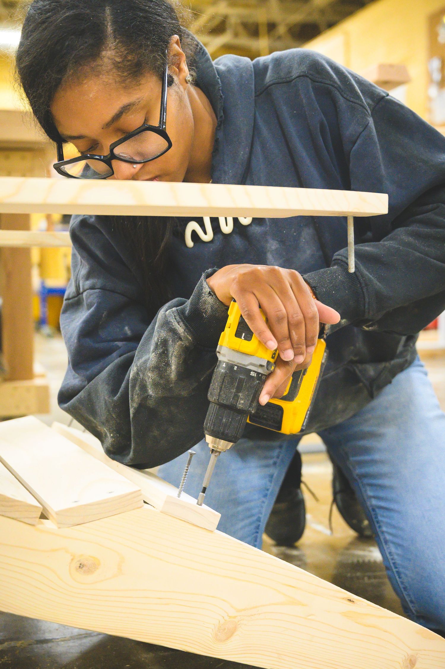 A woodworker wearing safety glasses uses a power drill to secure screws into a wooden plank.