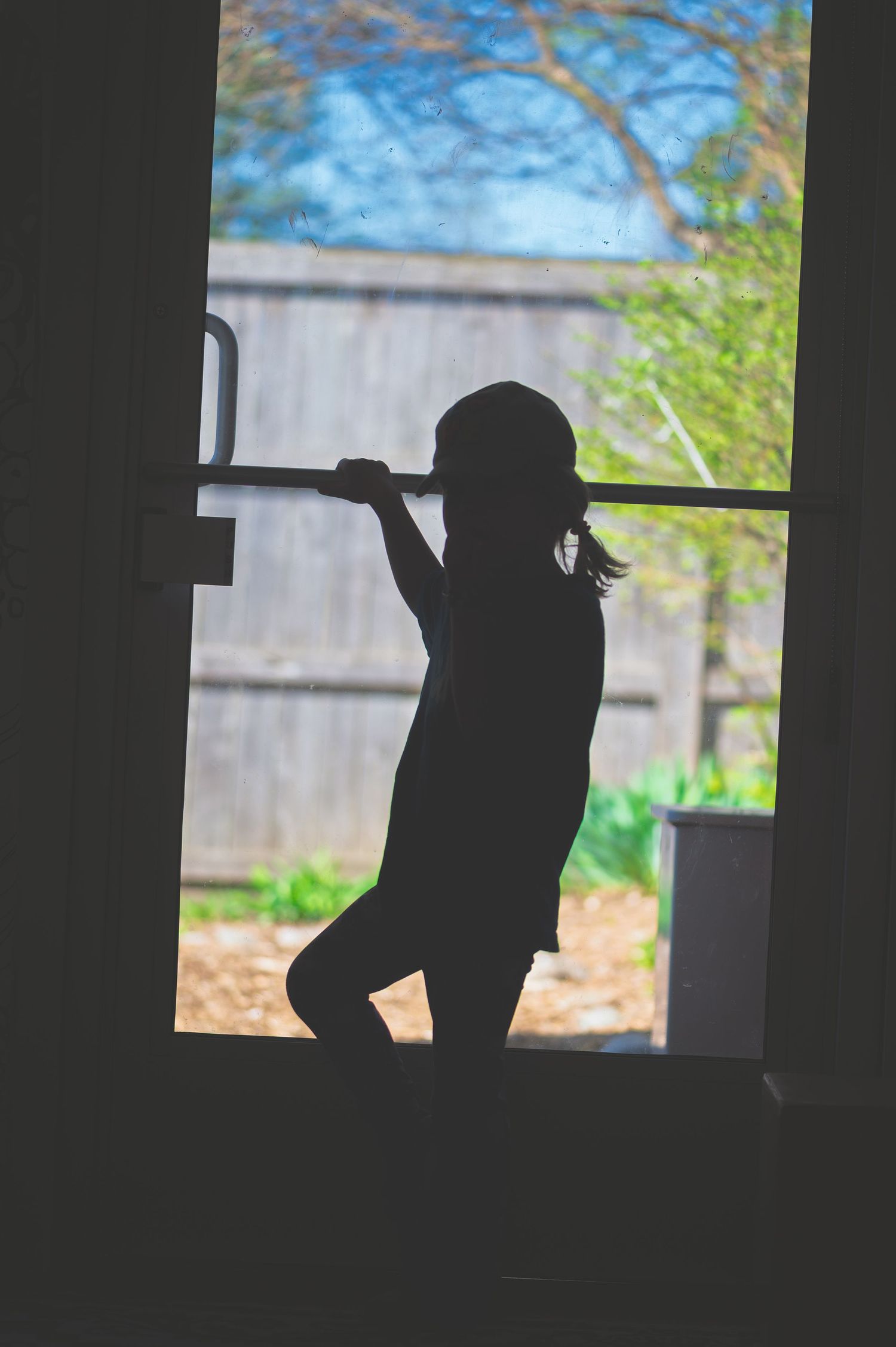 Silhouette of a child holding a bar in front of a bright window with a backyard view.