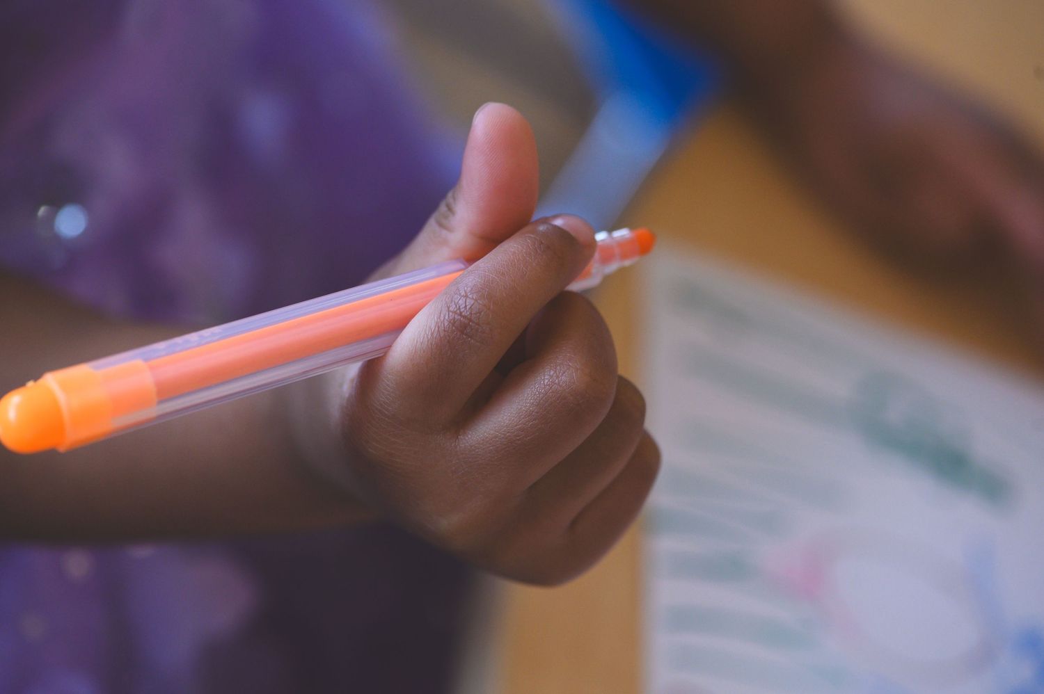 A child's hand holding an orange marker pen, ready to draw or write on paper.