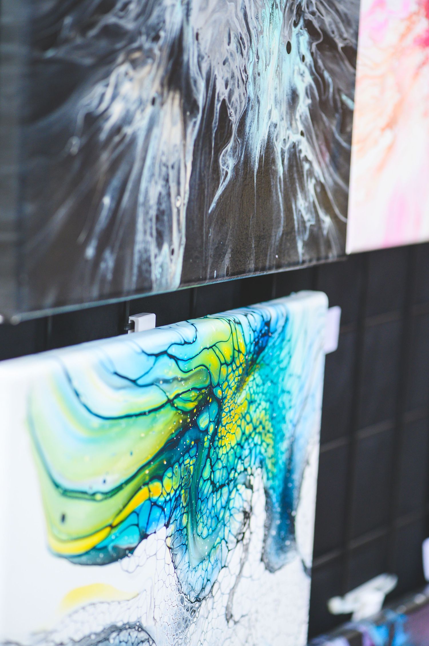 Vibrant fluid art paintings displayed outdoors, featuring swirling blue, green, and yellow abstract designs with marbled textures.