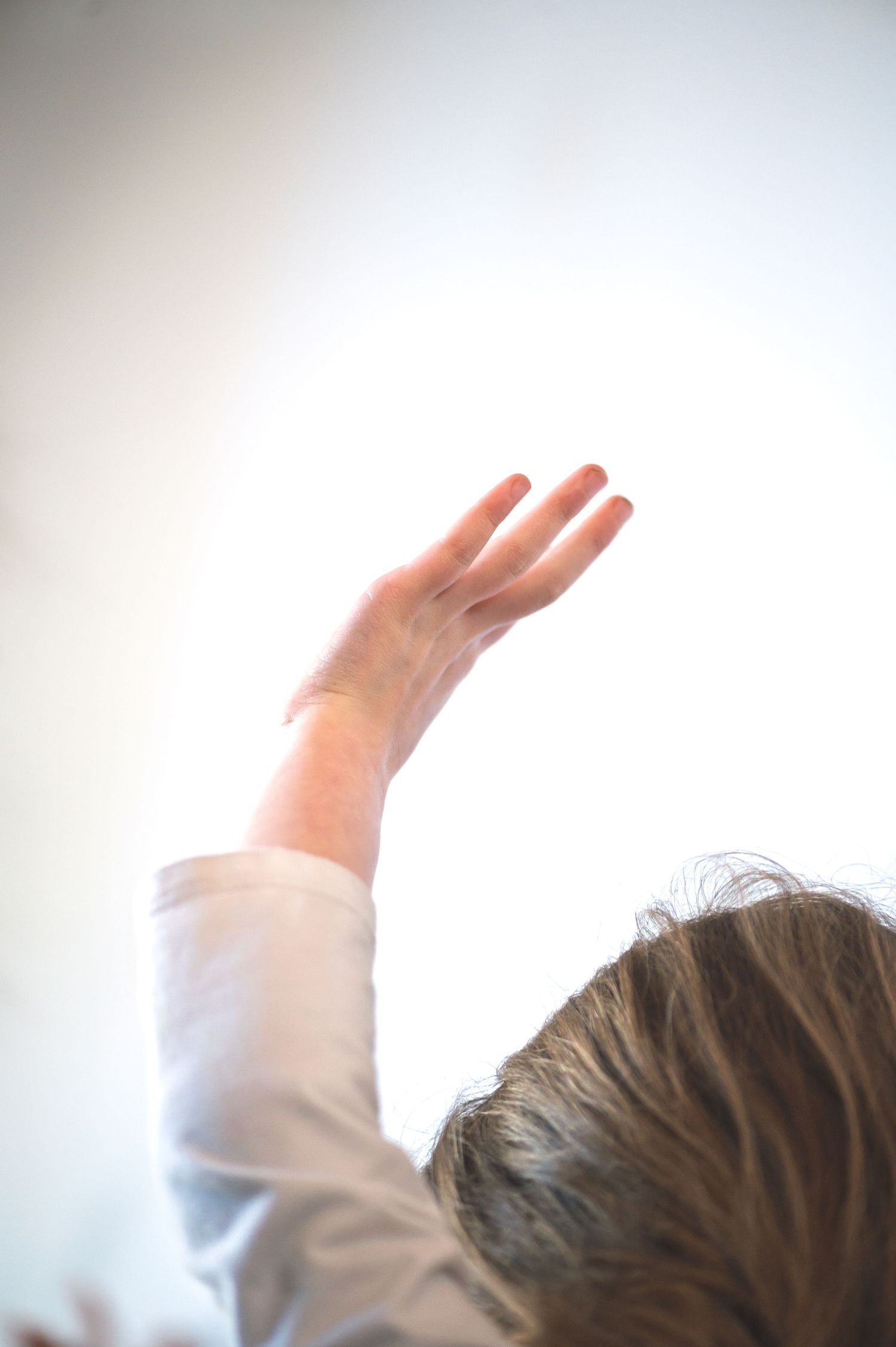 Person raising hand toward bright white light, viewed from behind, wearing light blue shirt.