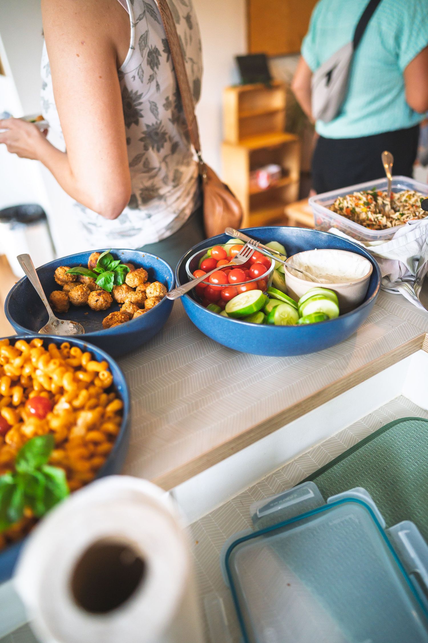 Blue bowls filled with fresh vegetables, tomatoes, cucumbers, roasted potatoes, and pasta salad on a wooden buffet table.