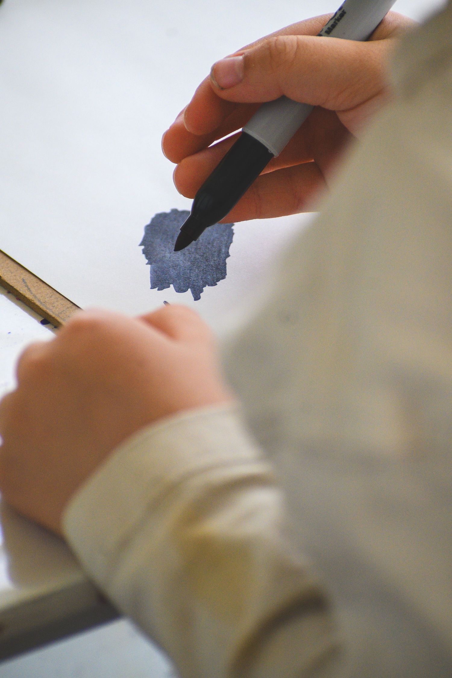 Child's hands drawing a blue ink blot on white paper with a black marker pen.