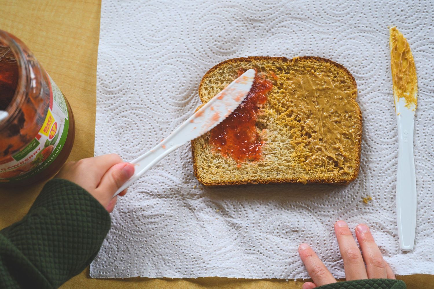 Child spreading peanut butter and jelly on whole wheat toast with a plastic knife on a paper towel.