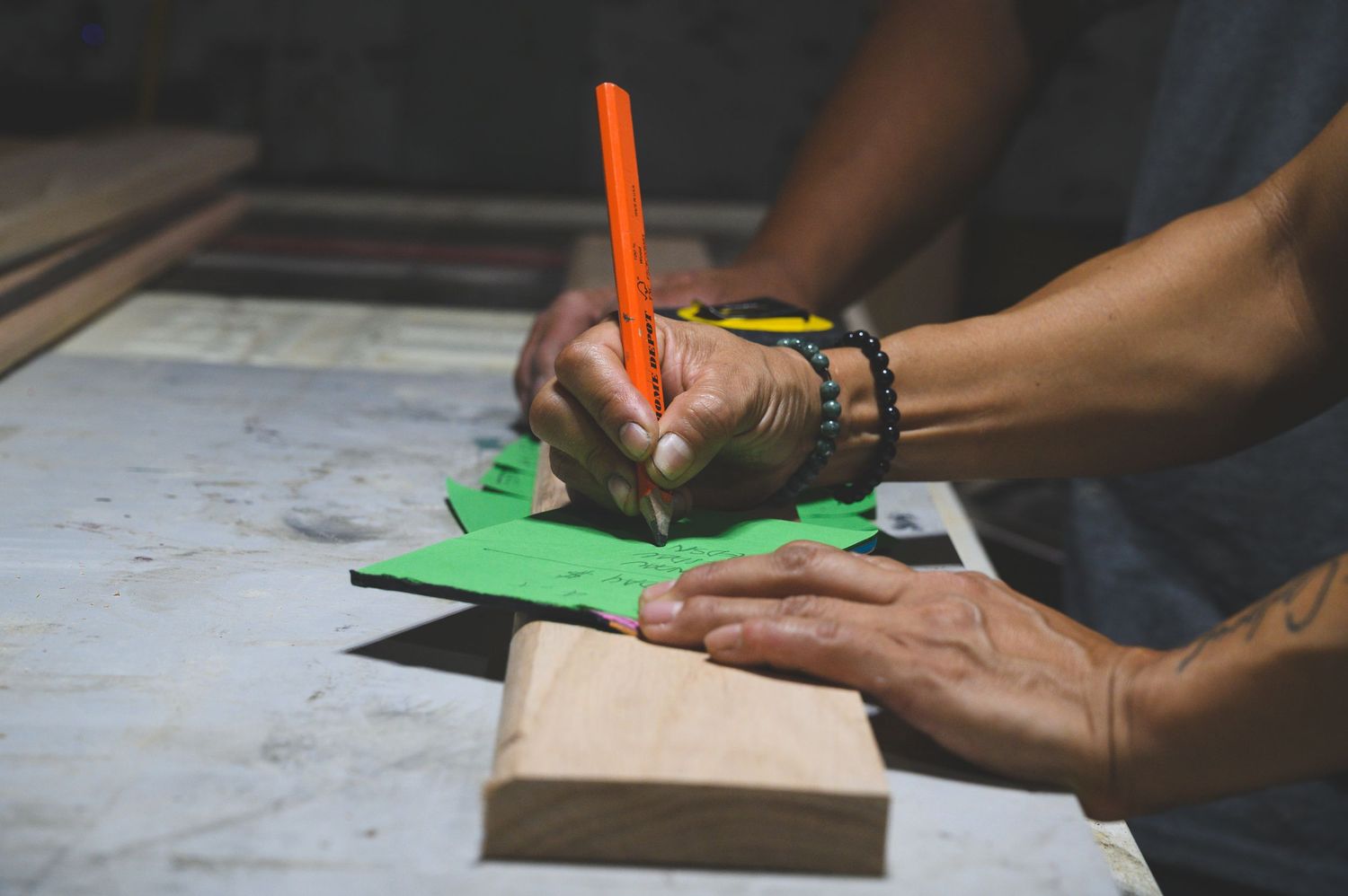 A craftsman traces a wooden block onto green paper using an orange pencil, wearing beaded bracelets on his wrist.