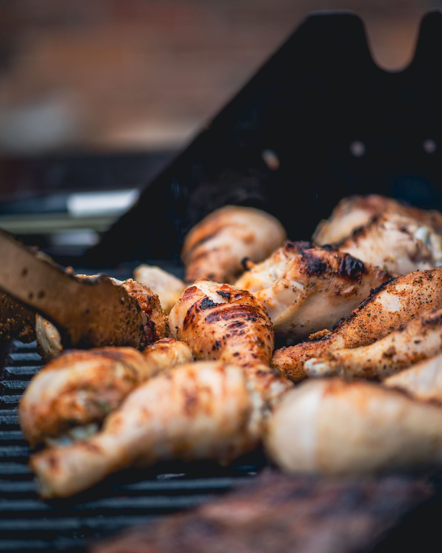 Seasoned chicken pieces grilling on a barbecue grill with tongs, showing golden-brown char marks.