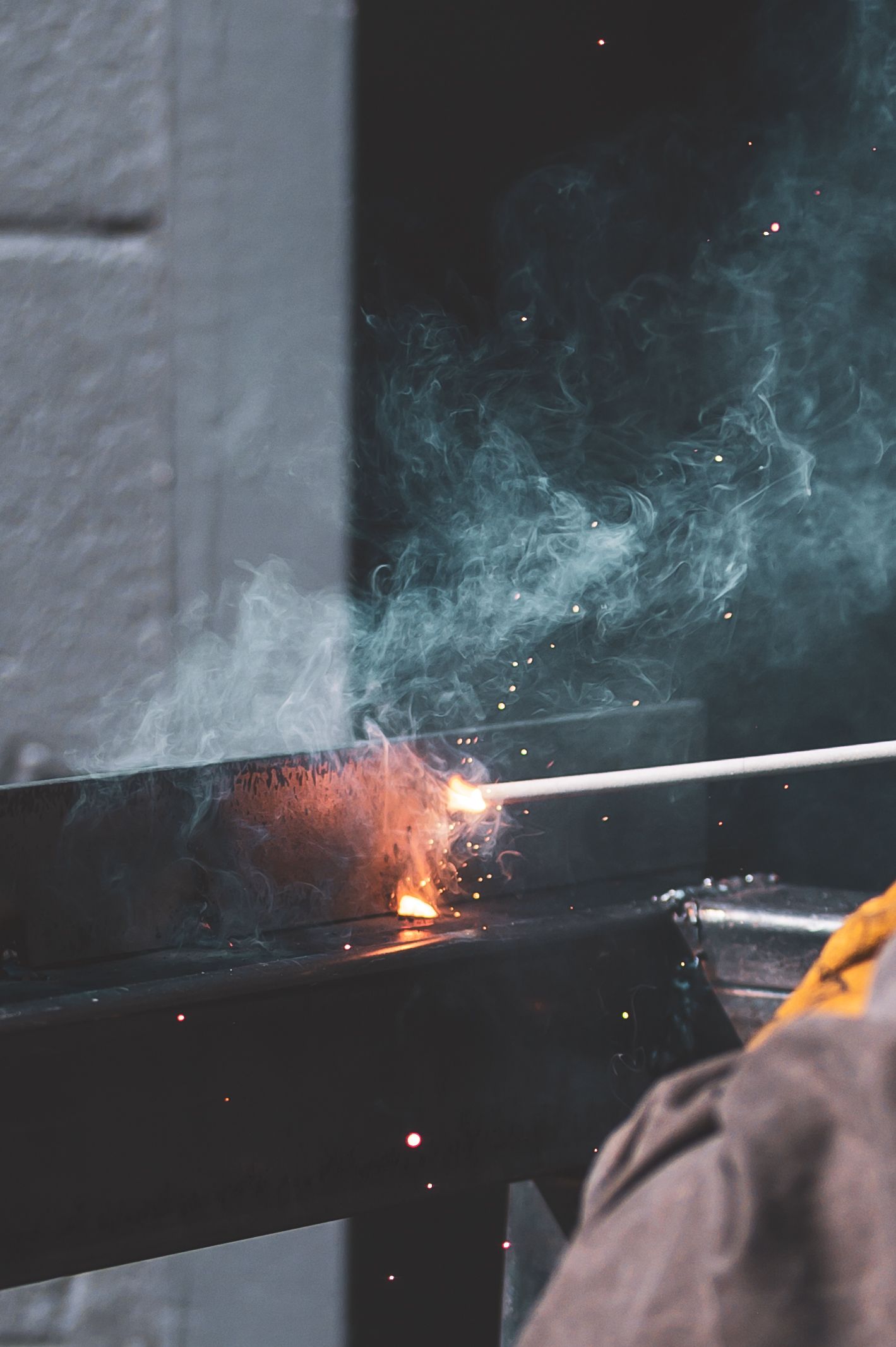 Close-up of a welder using a welding torch on metal, with bright sparks and smoke rising in a dark workshop.