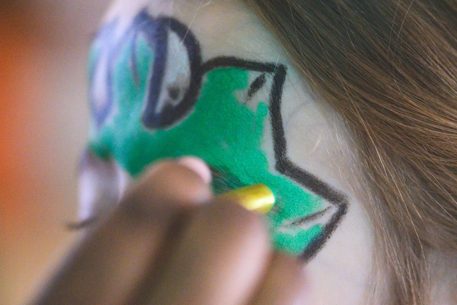 Close-up of green frog face paint being applied to a child's cheek with a yellow sponge.